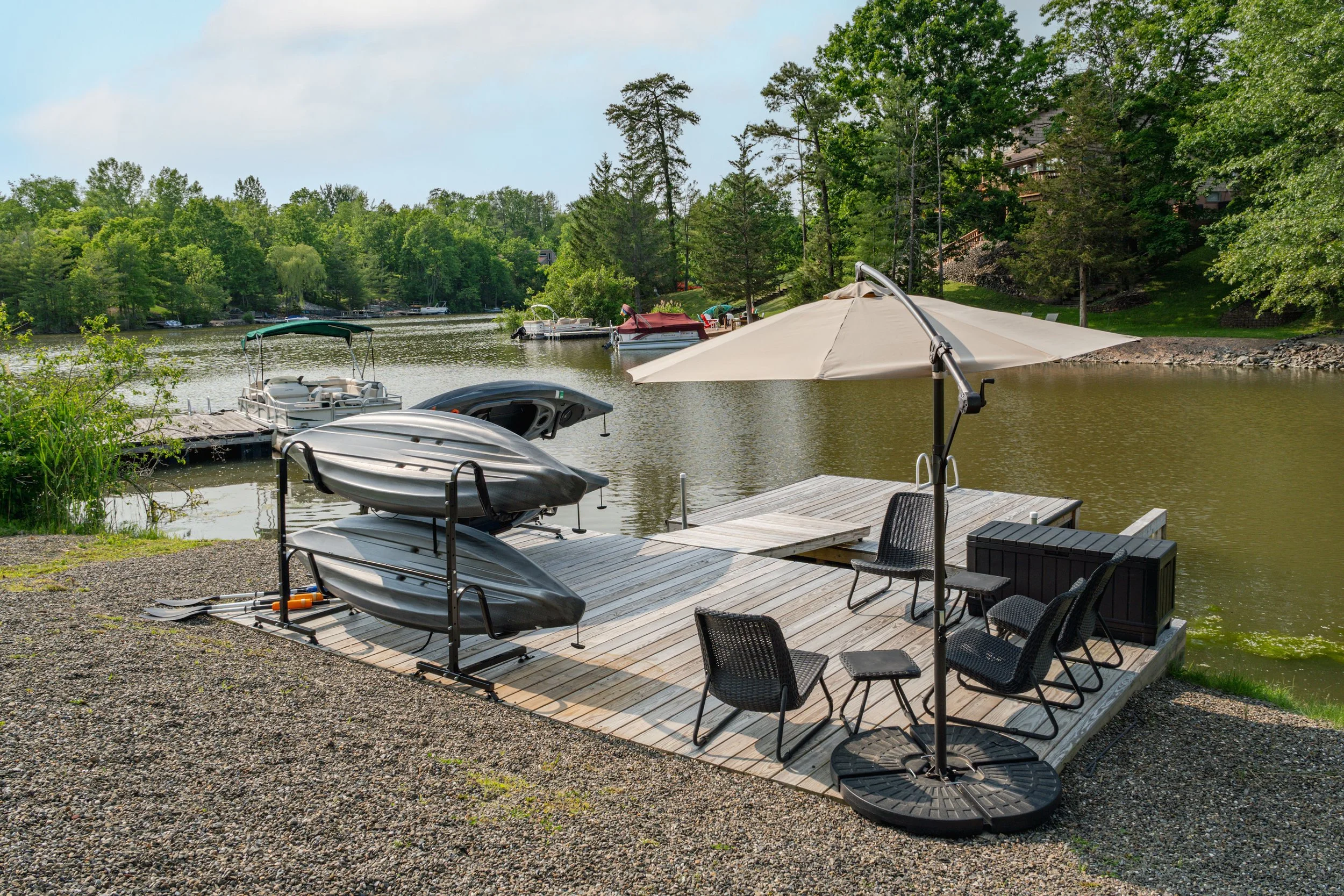 A lakeside dock with stacked kayaks, black chairs, a large umbrella, and boats on the water with trees and houses in the background.