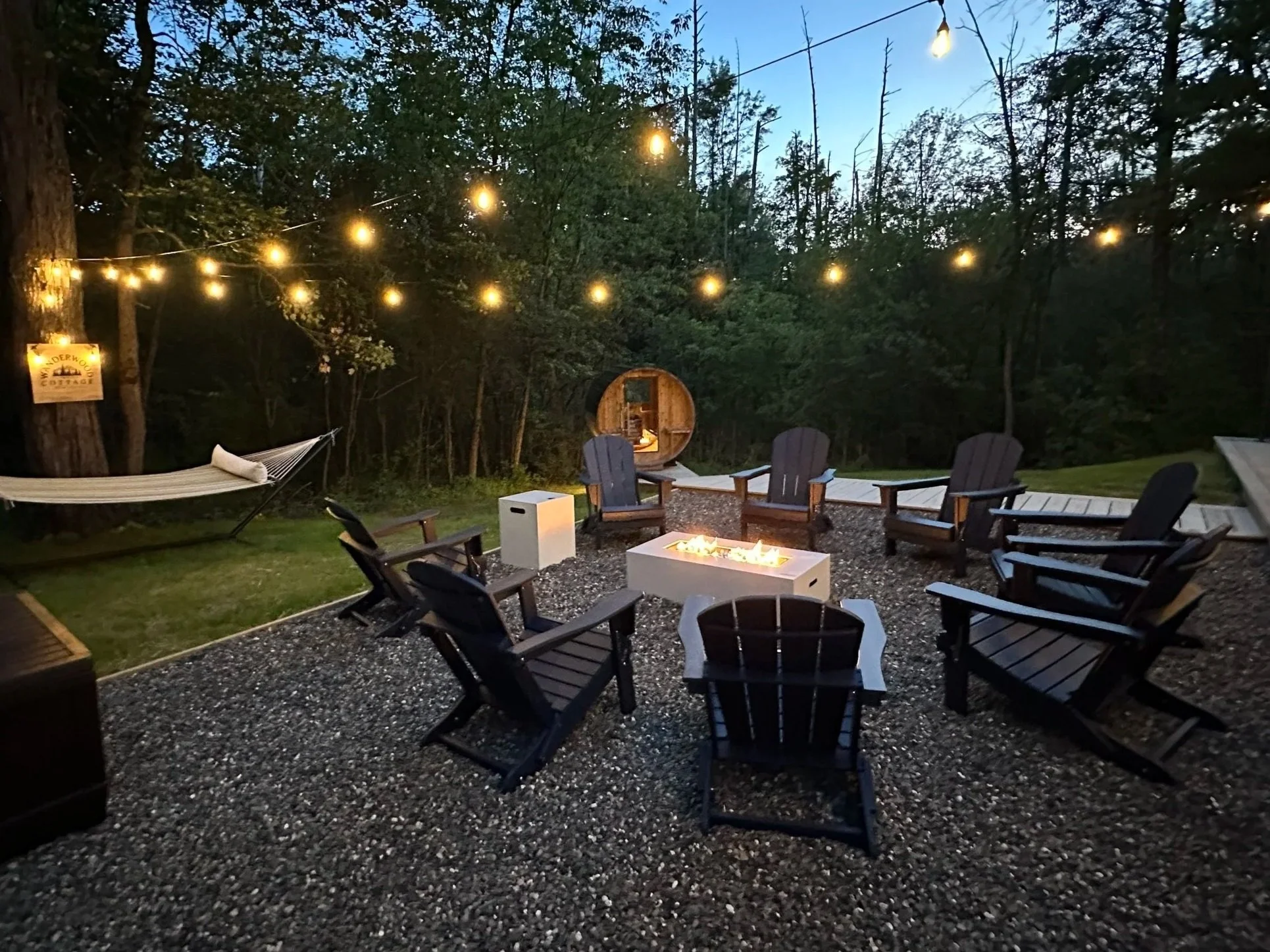 Outdoor fire pit with chairs arranged around it, string lights overhead, hammock on the left, surrounded by trees at dusk.