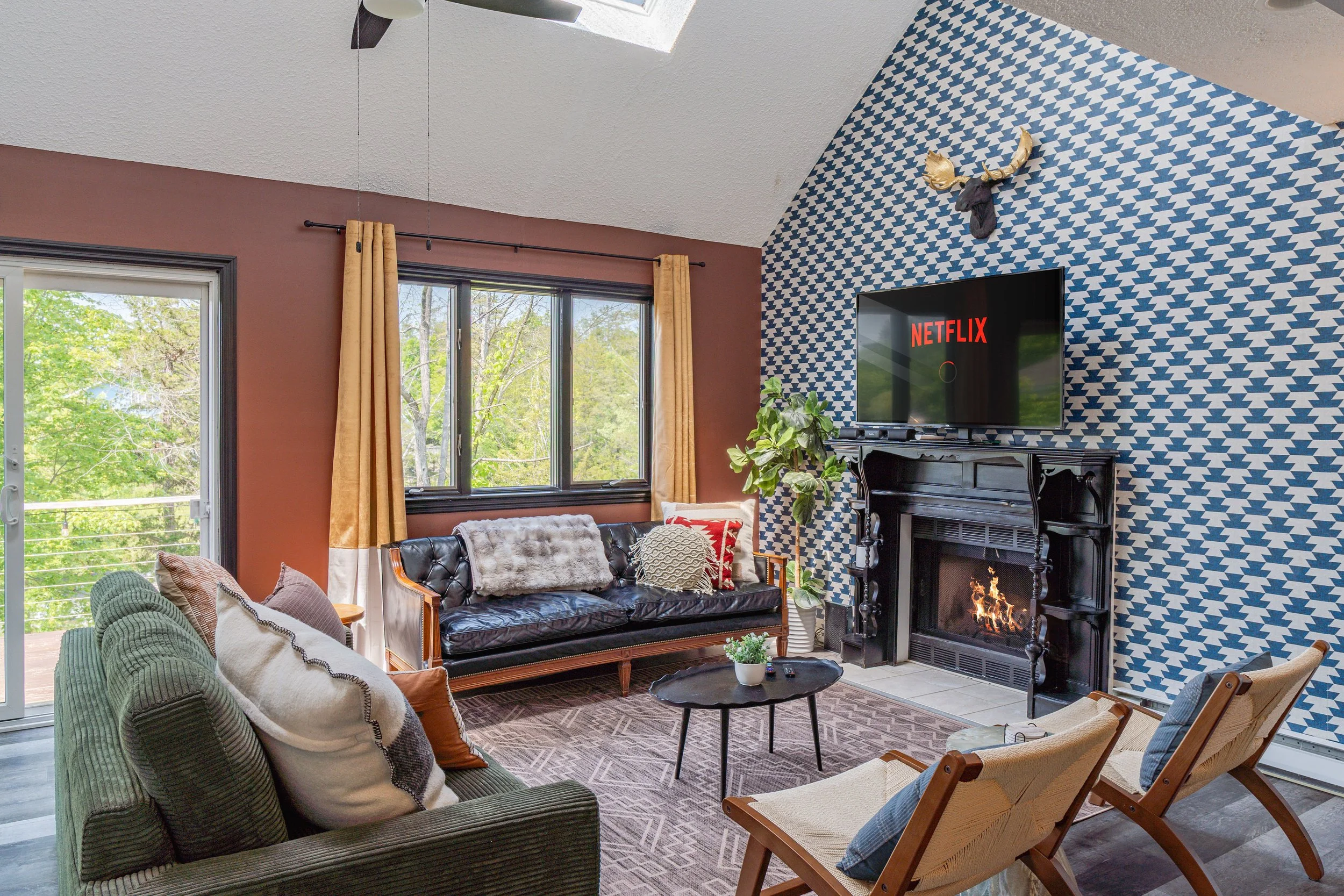 Cozy living room with patterned blue and white accent wall, black fireplace, wall-mounted TV displaying Netflix, large window with beige curtains, sliding glass door, and furniture including a green sofa, a black leather sofa with pillows, and two wooden chairs, with a small round black coffee table and a decorative moose head on the wall.