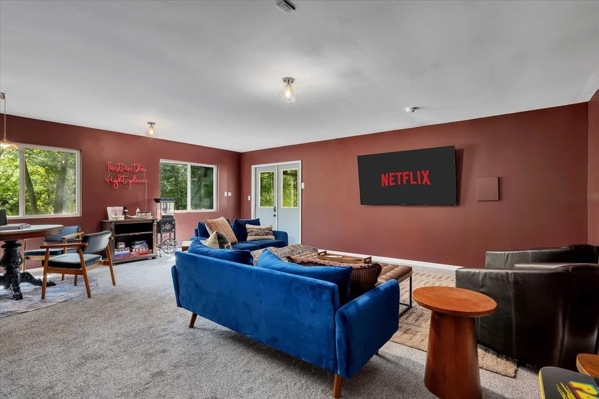Living room with maroon accent wall, two large windows, a door, a blue sofa, black armchair, small wooden side table, and a flat-screen Netflix TV mounted on the wall.