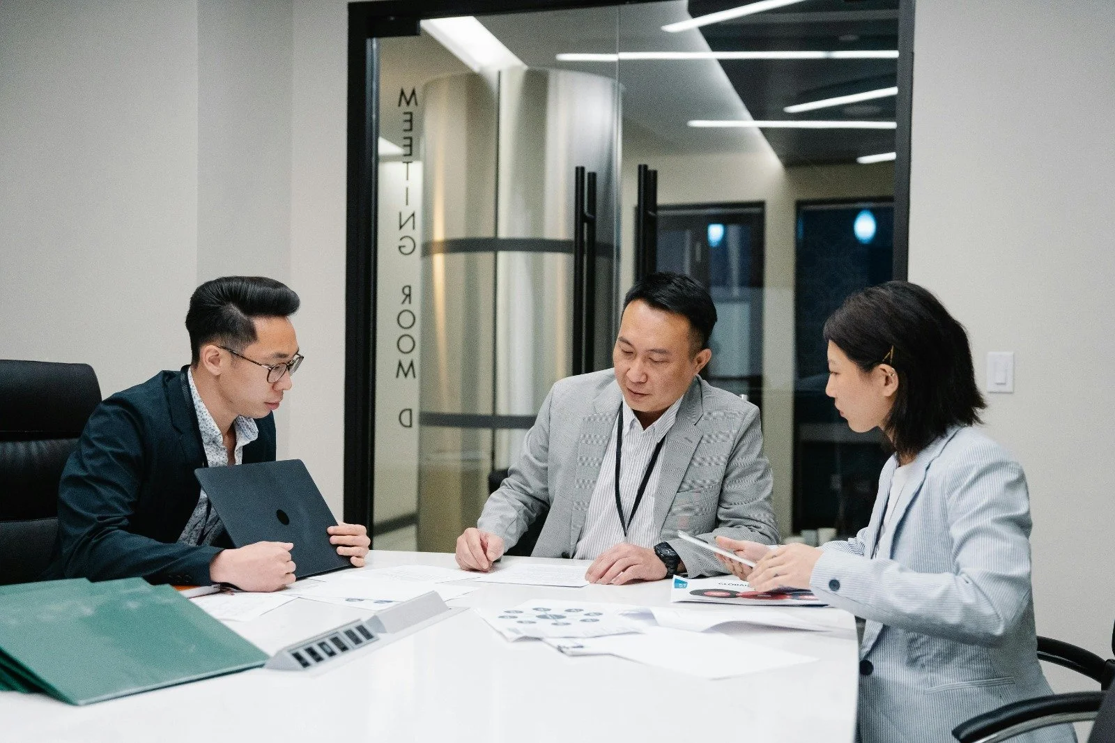 Three business professionals sitting at a conference table in a modern office discussing documents and digital devices.