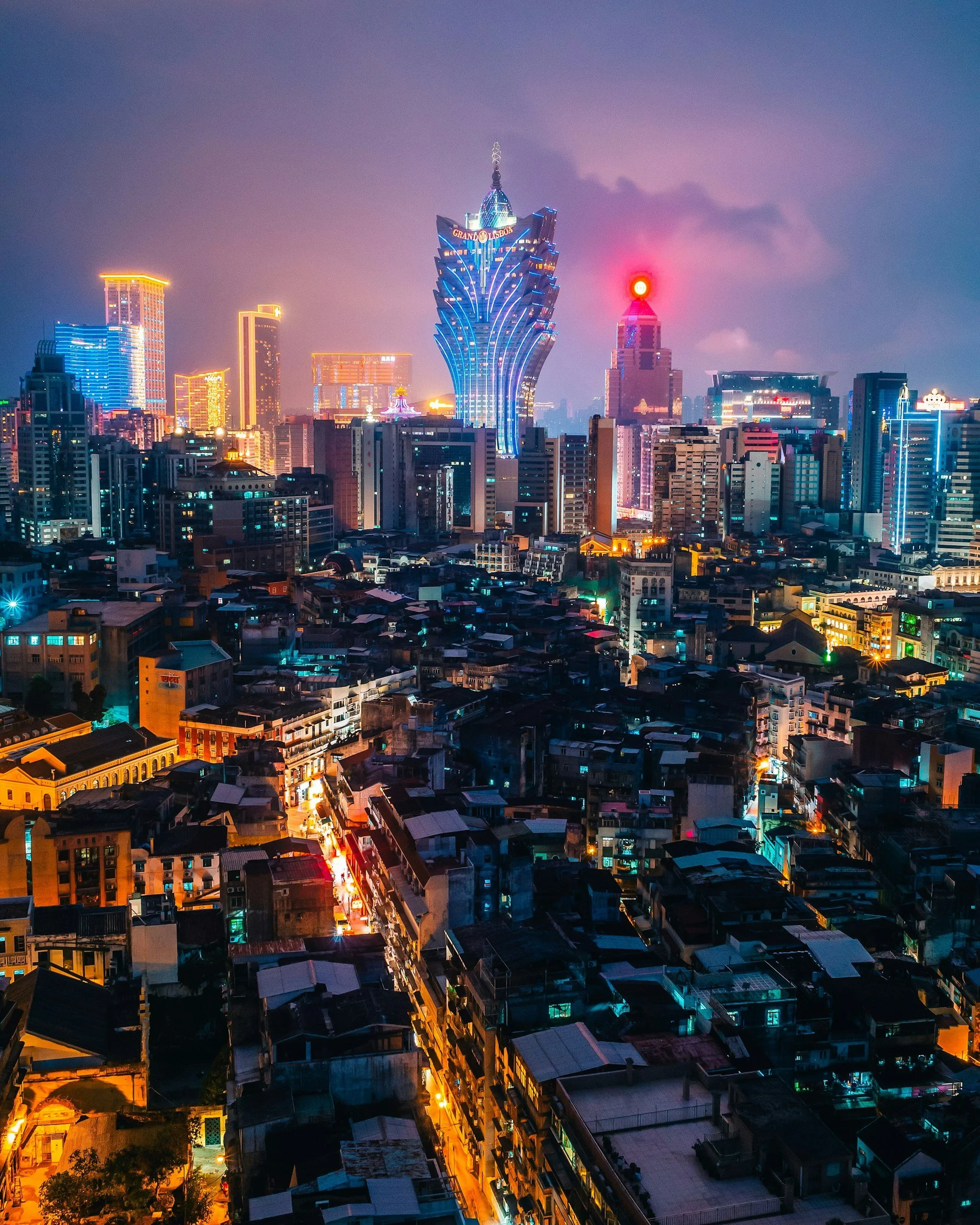 Night city skyline with illuminated skyscrapers, including the Grand Lisboa hotel with its distinctive design, and colorful buildings with some neon lighting, under a partly cloudy sky.