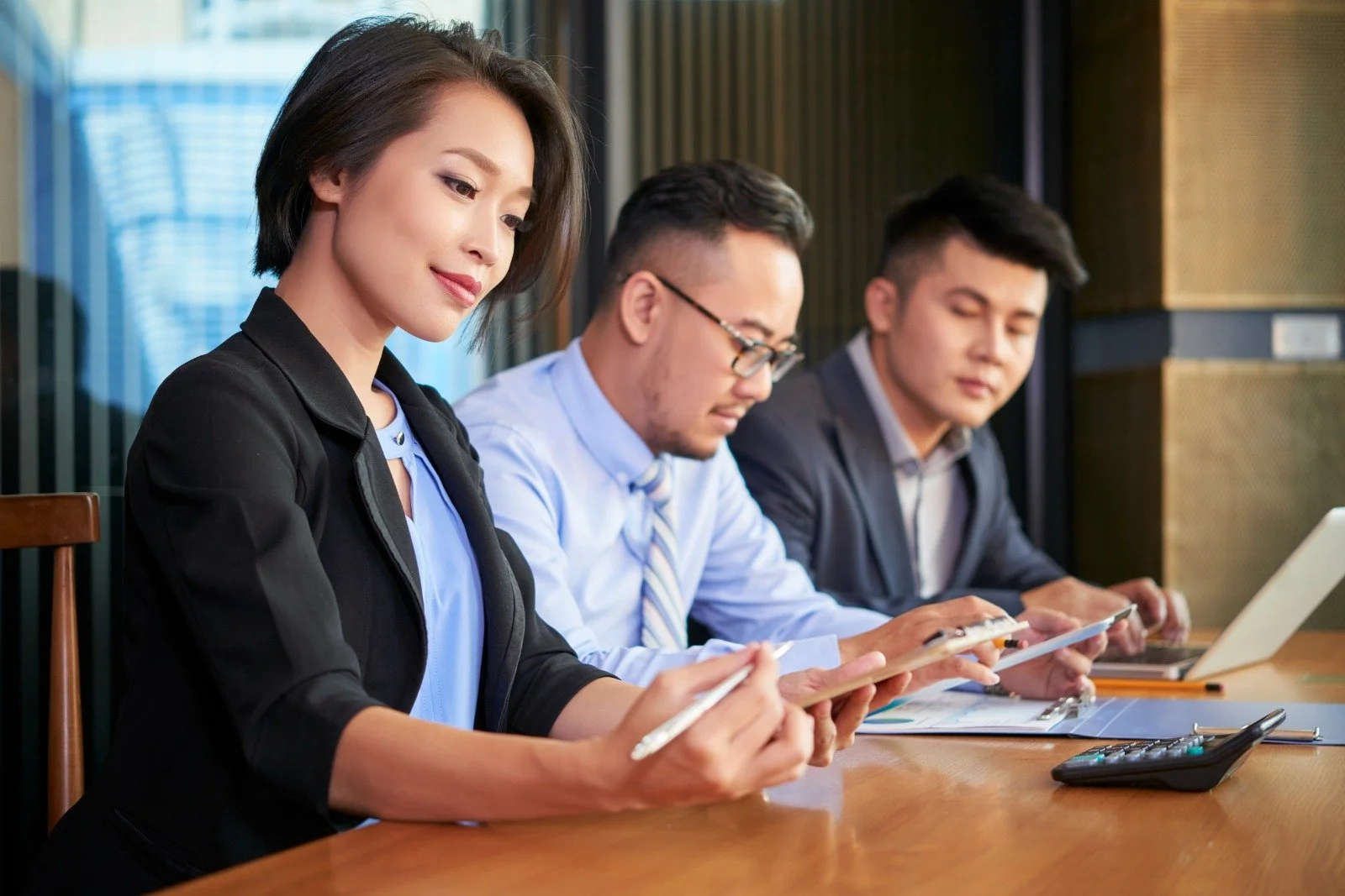 Three people sitting at a conference table in a business meeting, using tablets and laptops.