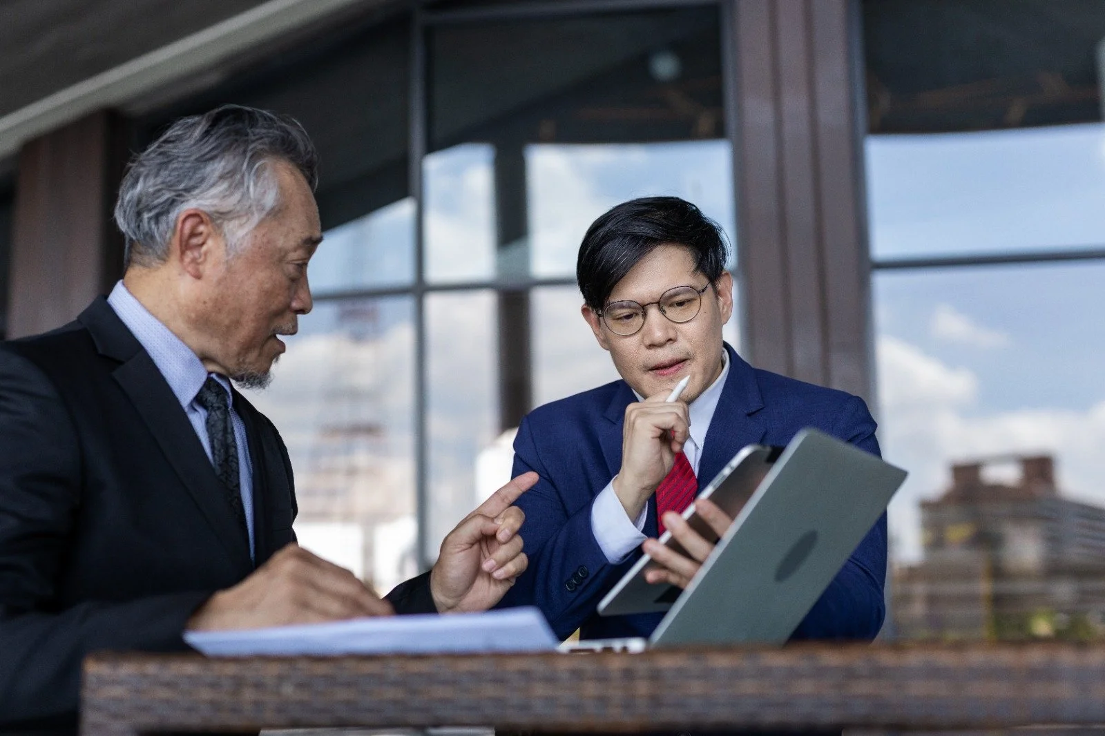 Two businessmen in suits having a discussion, looking at a tablet together indoors with large windows in the background.