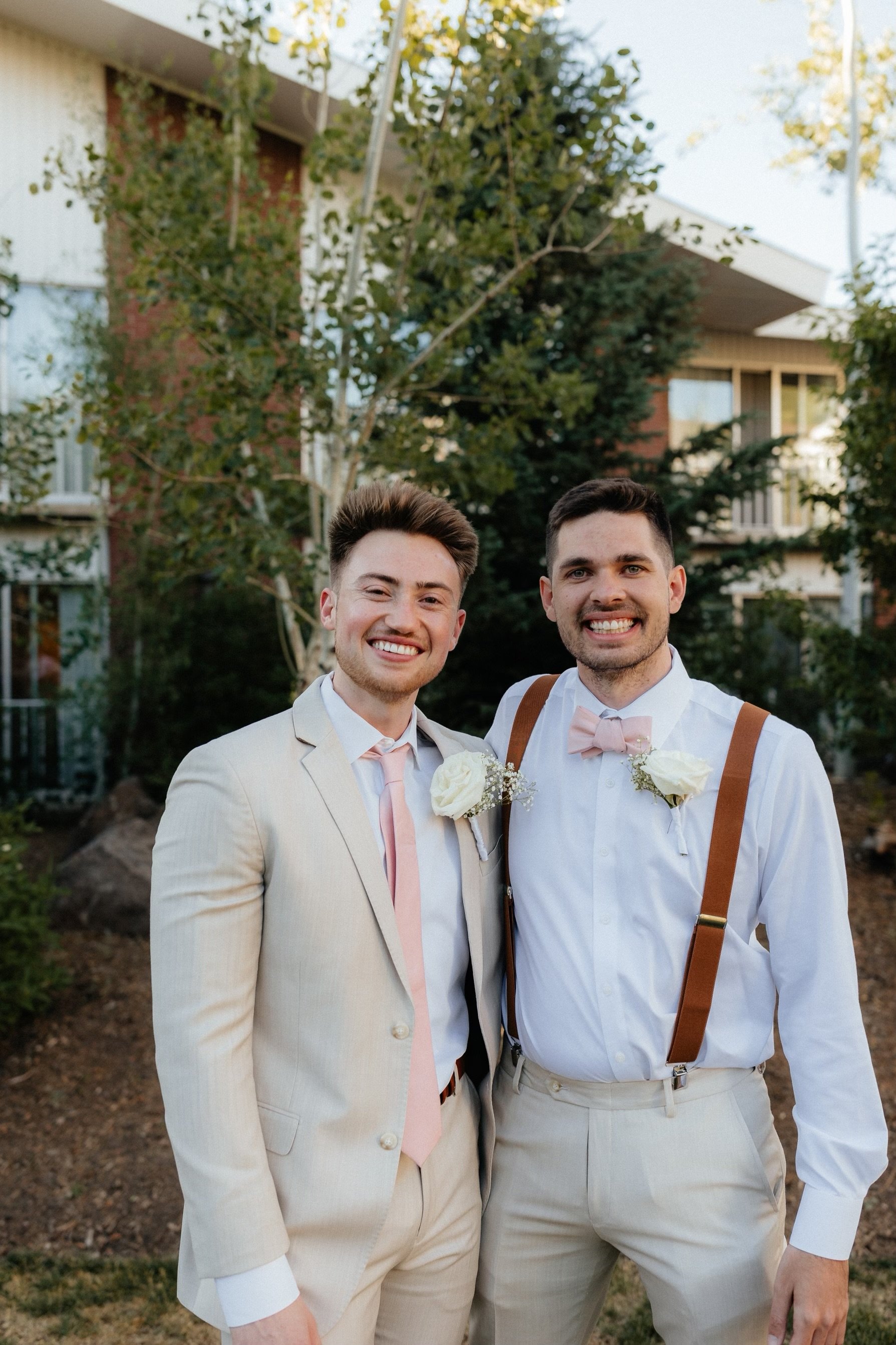 Two men dressed in wedding attire, smiling for a photo outdoors with trees and buildings in the background.