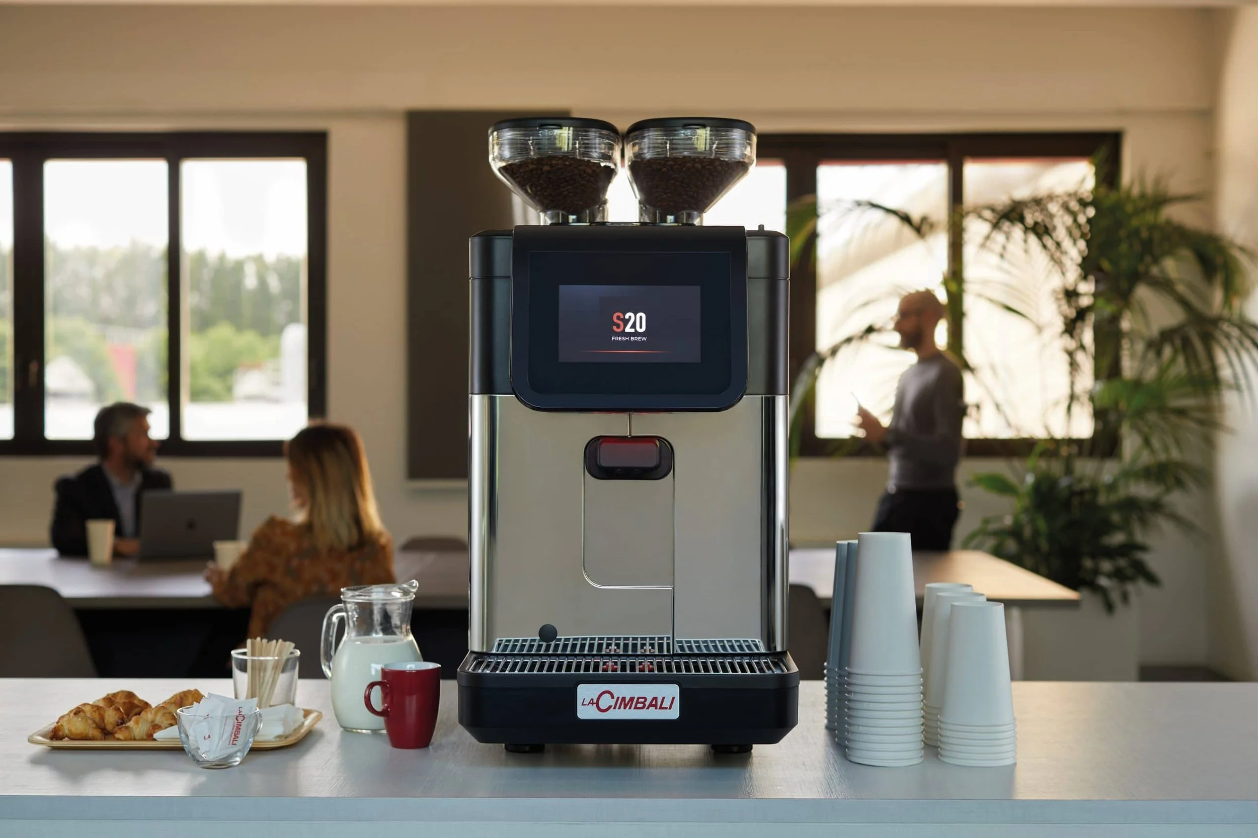 Coffee machine with beans in hoppers, cups, pastries, and beverages on a counter in a modern office break room with people working and socializing in the background.