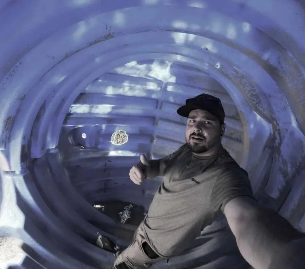 A man taking a selfie inside a large, blue plastic pipe with a textured surface, giving a thumbs-up gesture.