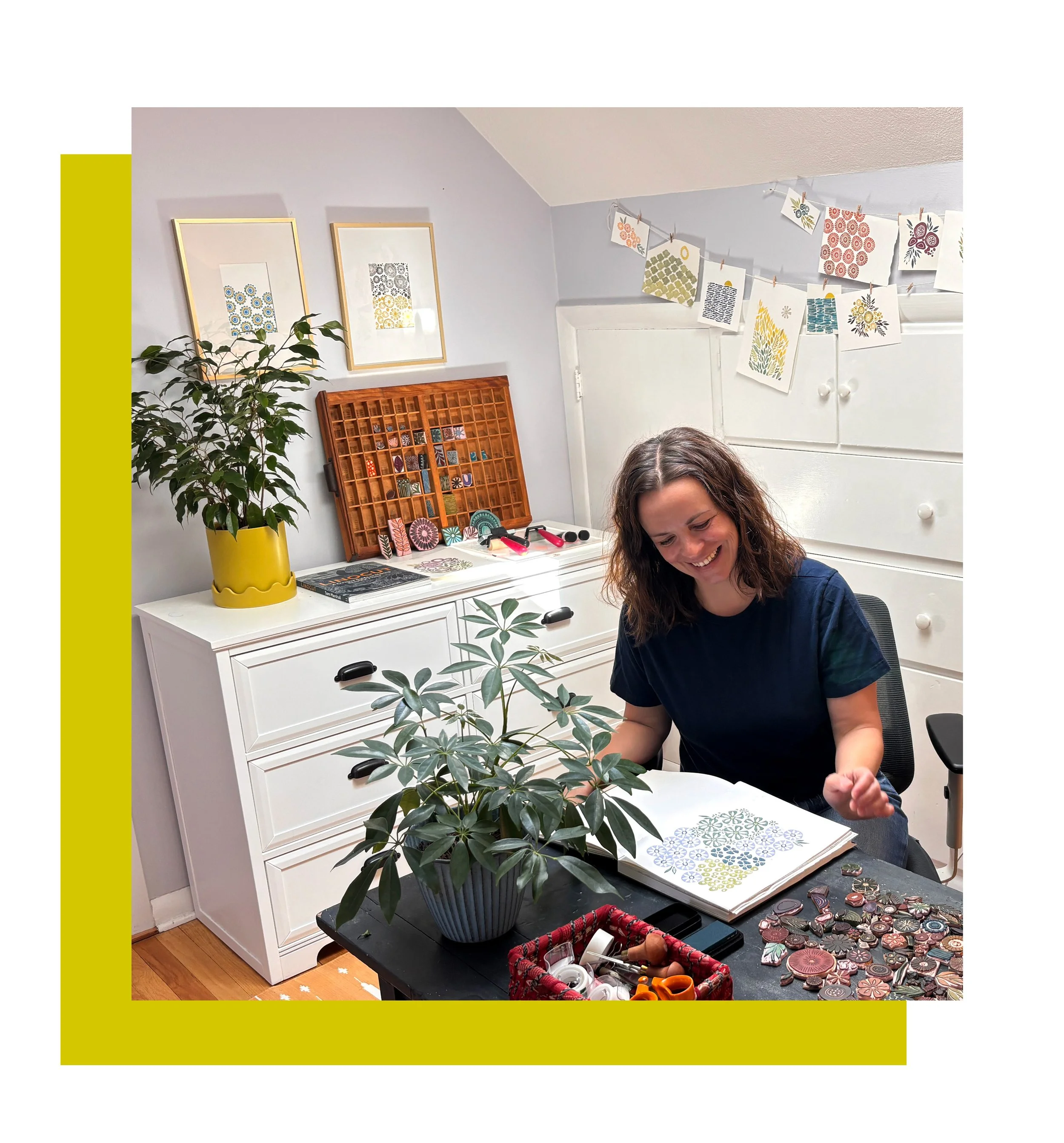 A woman sitting at a table with a large potted plant, smiling as she looks at an open book with patterns. The table has craft supplies including buttons and scissors. Behind her, there is a white dresser, artwork on the wall, a yellow pot with a leafy plant, and a display of crafted paper on a string.