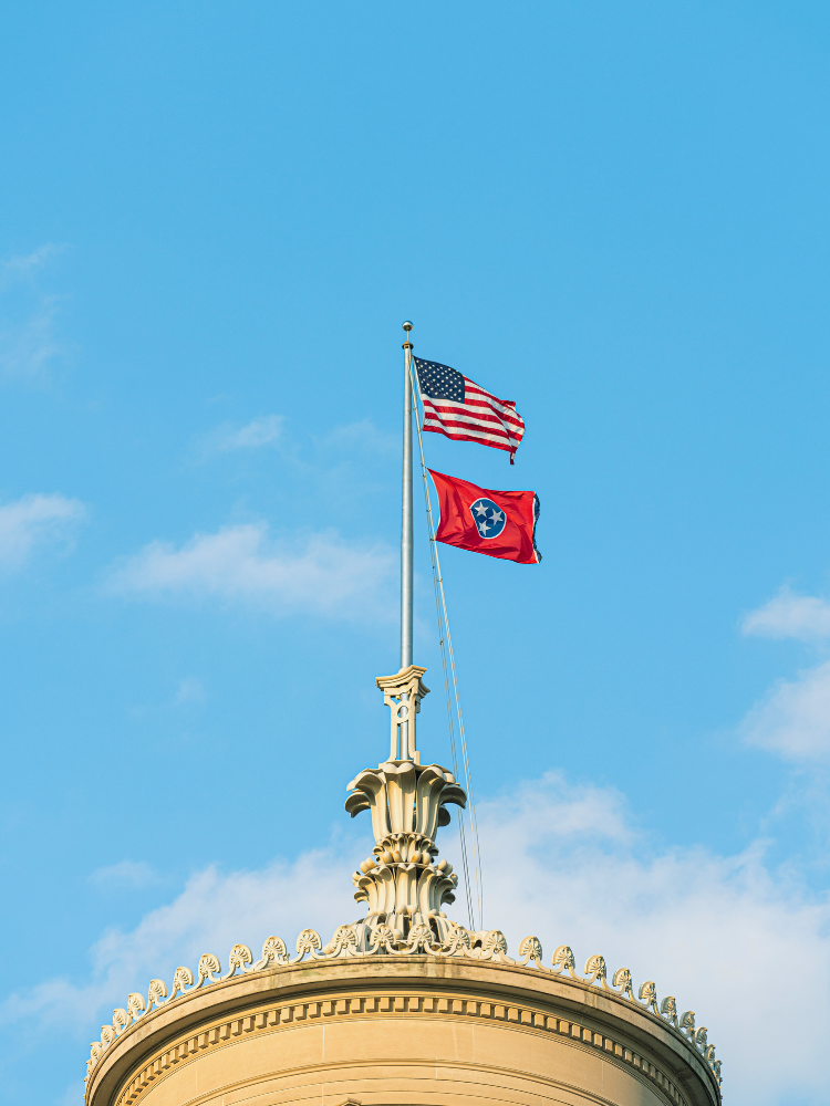 American and Tennessee flags flying atop a building with ornate architecture against a blue sky.