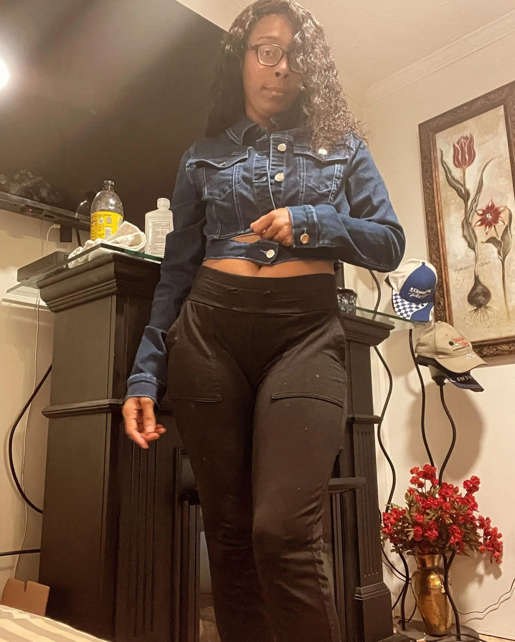 A woman with curly hair, glasses, wearing a denim jacket and black pants, standing in a room with a fireplace, framed art, hats, and a vase with red flowers.