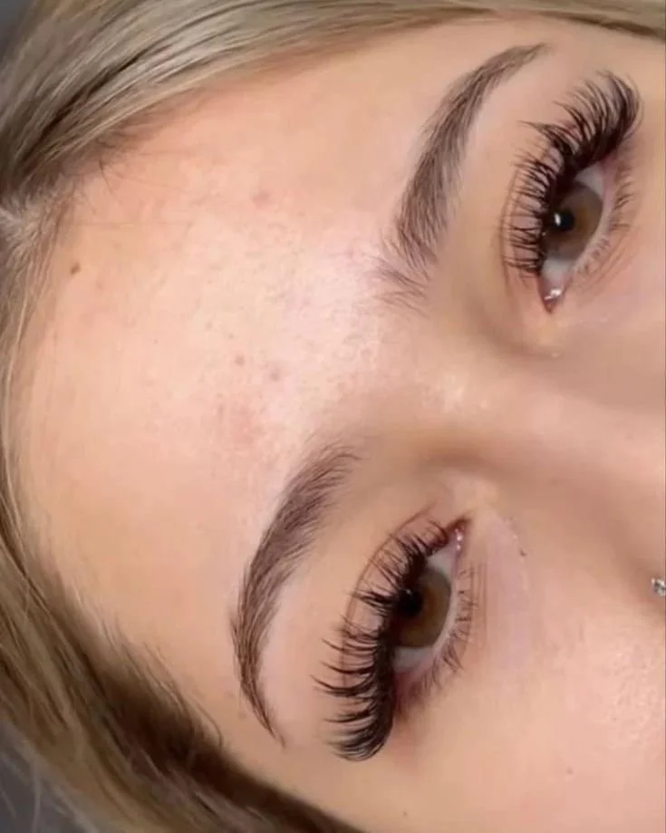 Close-up of a woman's face showing her eyes, eyebrows, and part of her cheek. She has long eyelashes, well-shaped eyebrows, and a small nose piercing.