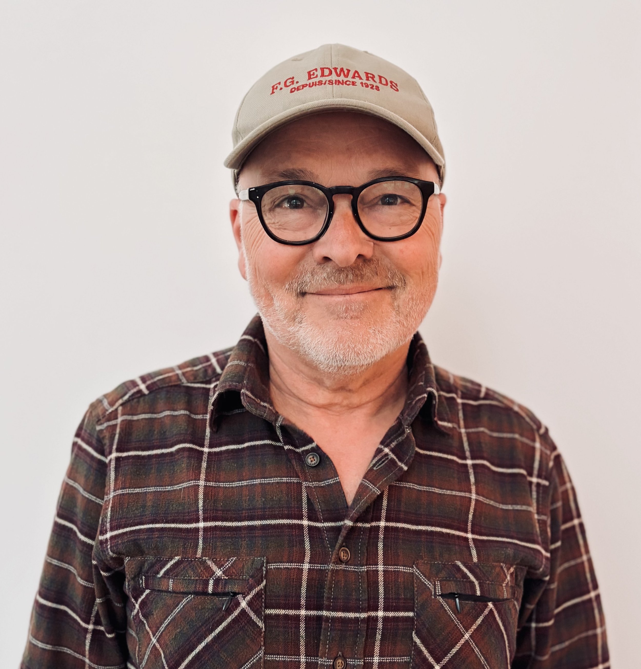 Un homme âgé souriant, porteur de lunettes noires, un chapeau beige avec inscription rouge et un vêtement à carreaux, se tient devant un mur blanc.