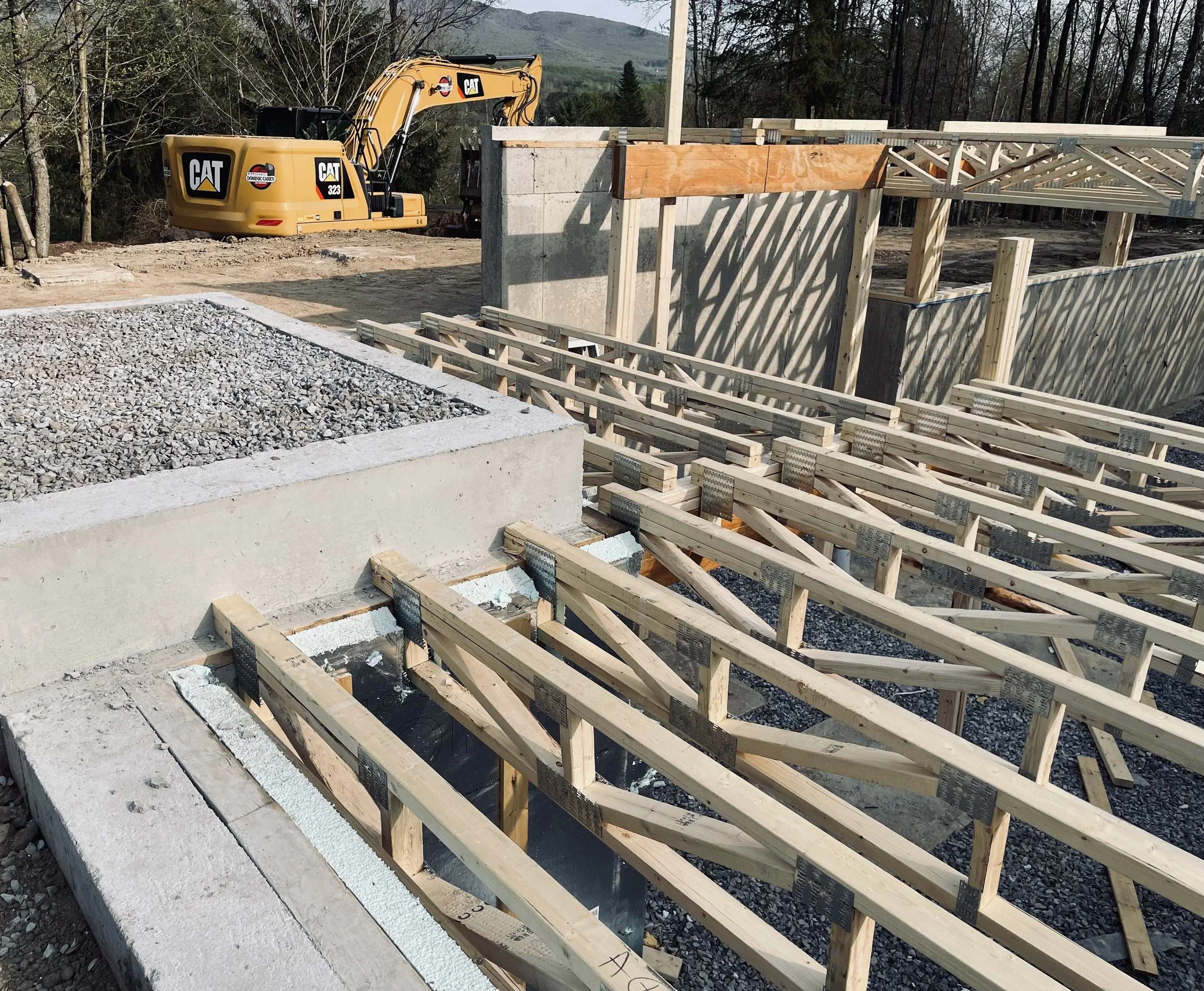 Construction de fondations avec ossature en bois, une excavatrice Caterpillar en arrière-plan, sur un site en plein air entouré d'arbres et de montagnes.