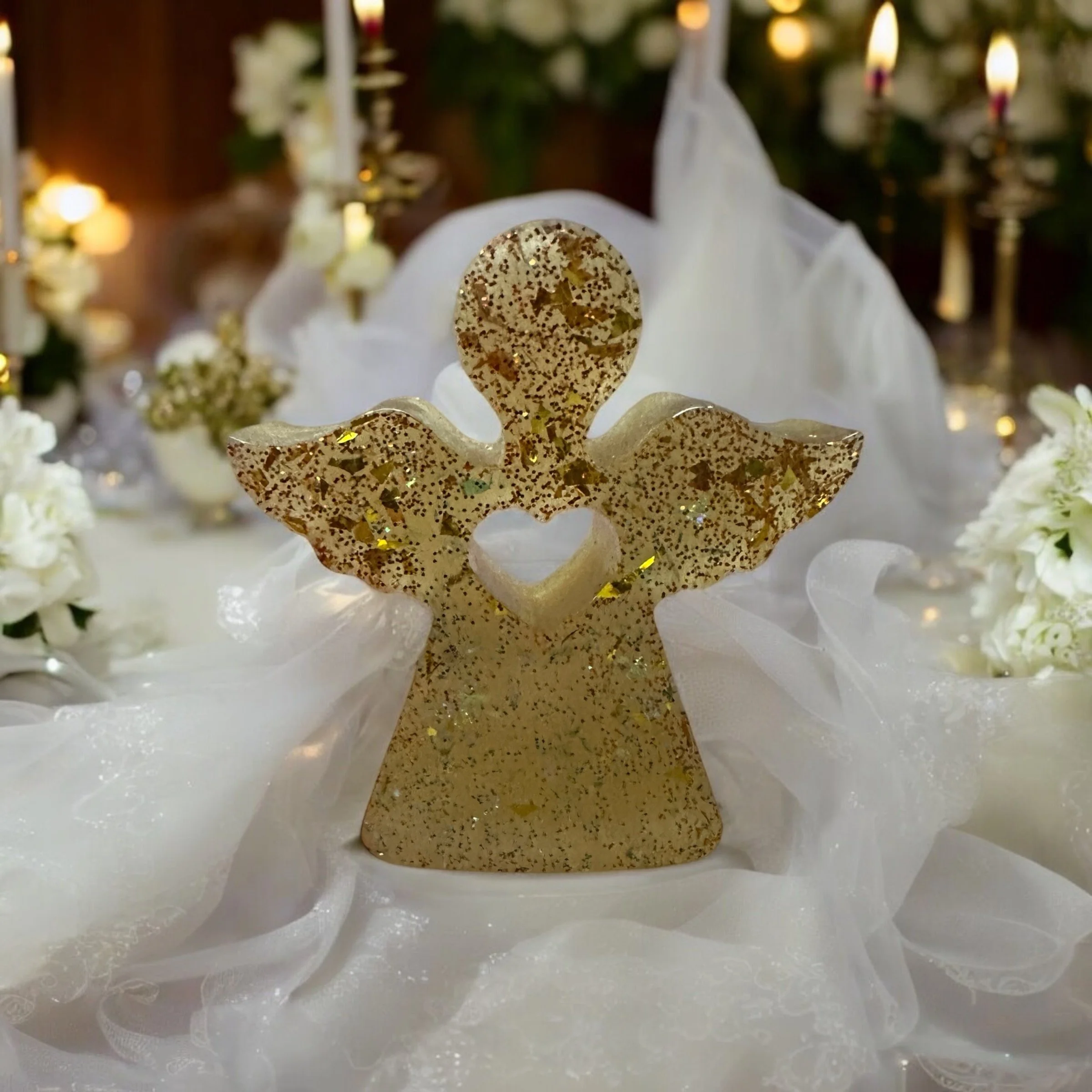 A golden angel figurine with glitter, holding a small heart, surrounded by white flowers and candles, on a white fabric tablecloth, with a softly lit background.