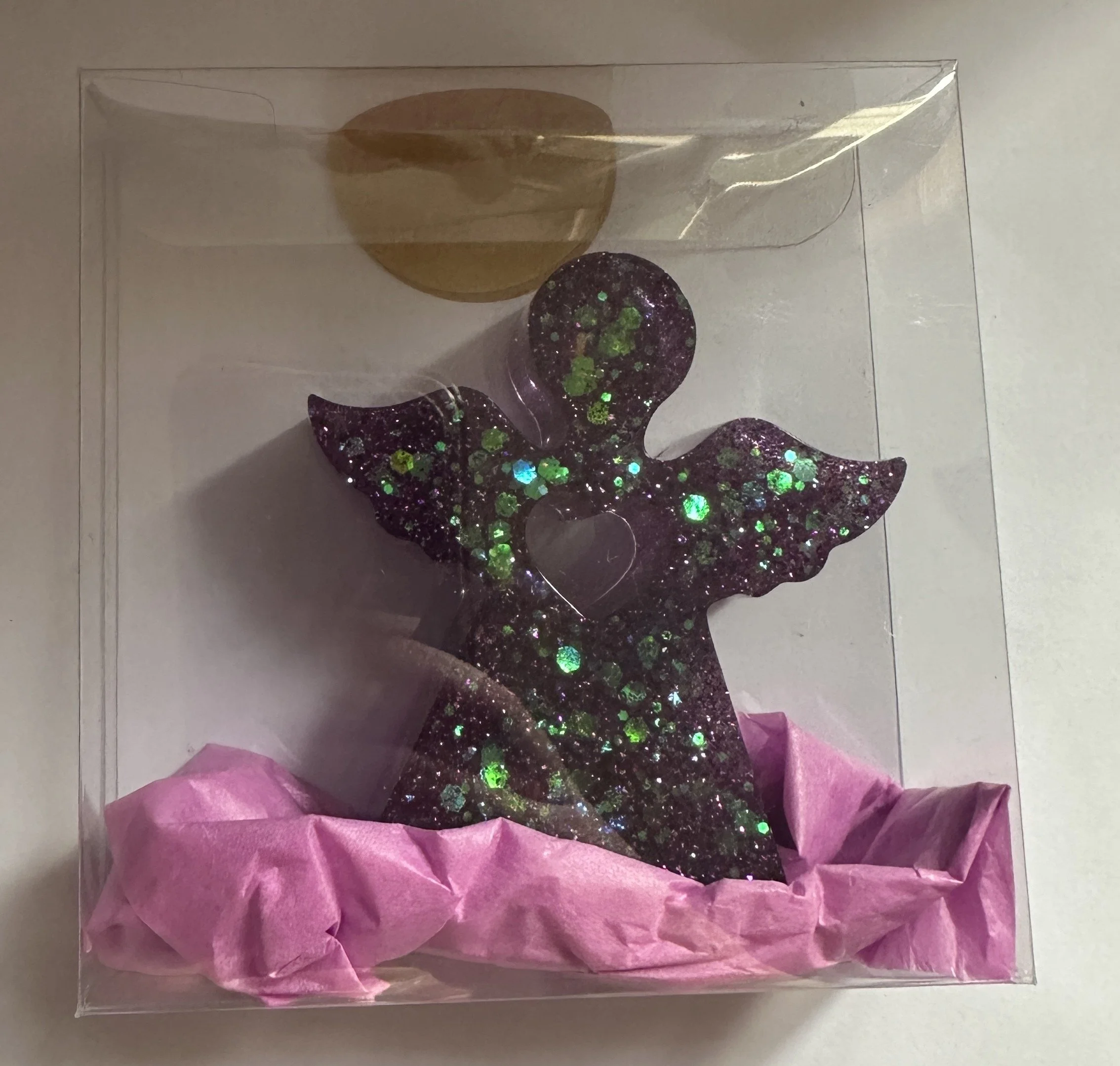 A glittery angel figure with wings and a heart-shaped cutout in the center, placed inside a clear display box with pink tissue paper at the bottom.