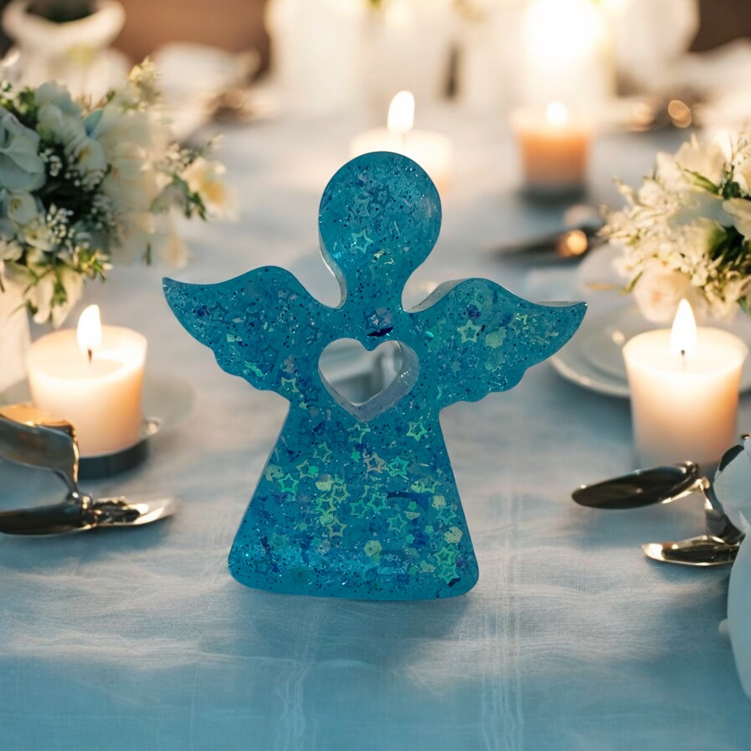 A blue decorative angel with a heart cutout in the chest, placed on a table with white tablecloth, surrounded by candles and floral arrangements.