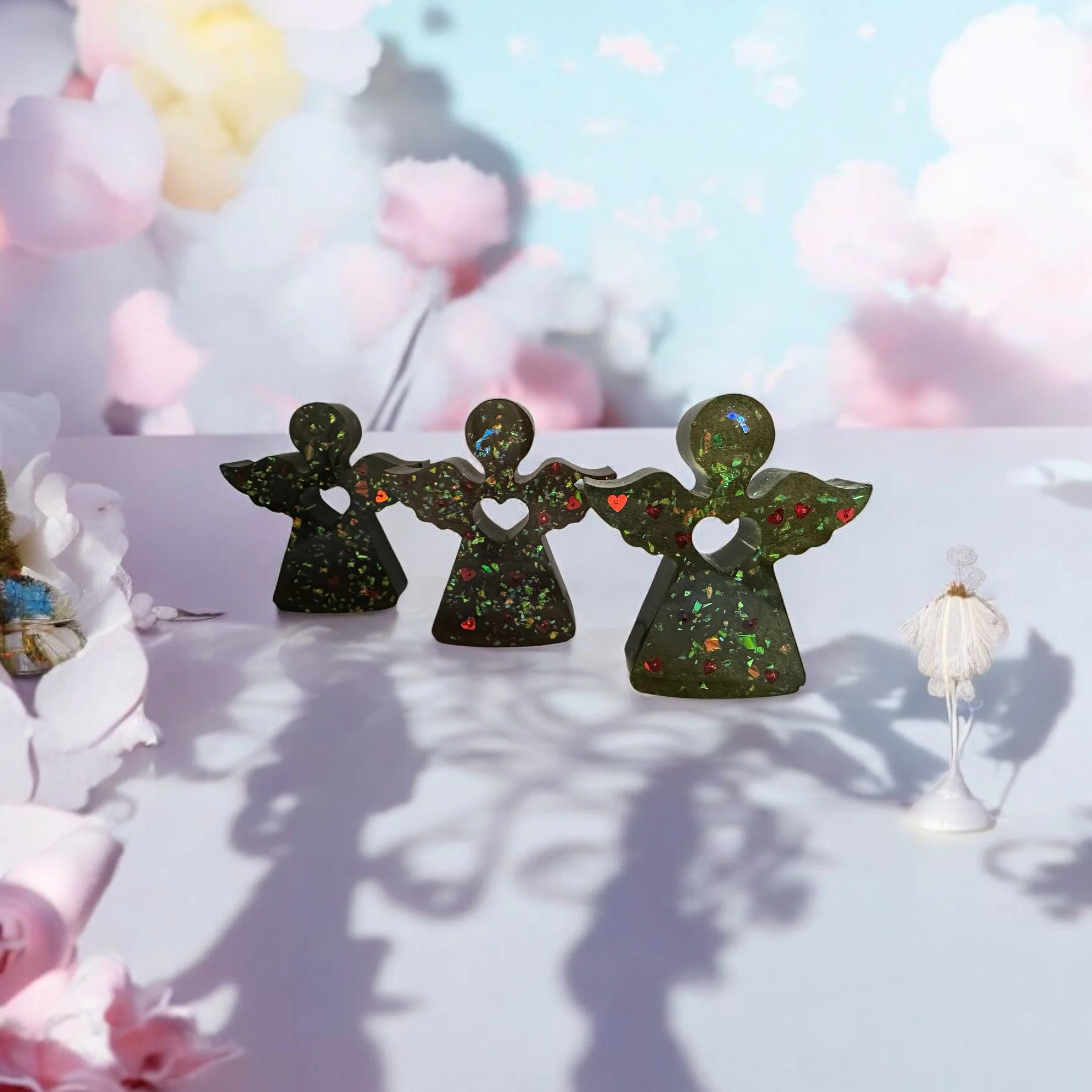 Three glass angel figurines with colorful glitter, standing on a white surface, with pink cloud background and soft lighting casting shadows.