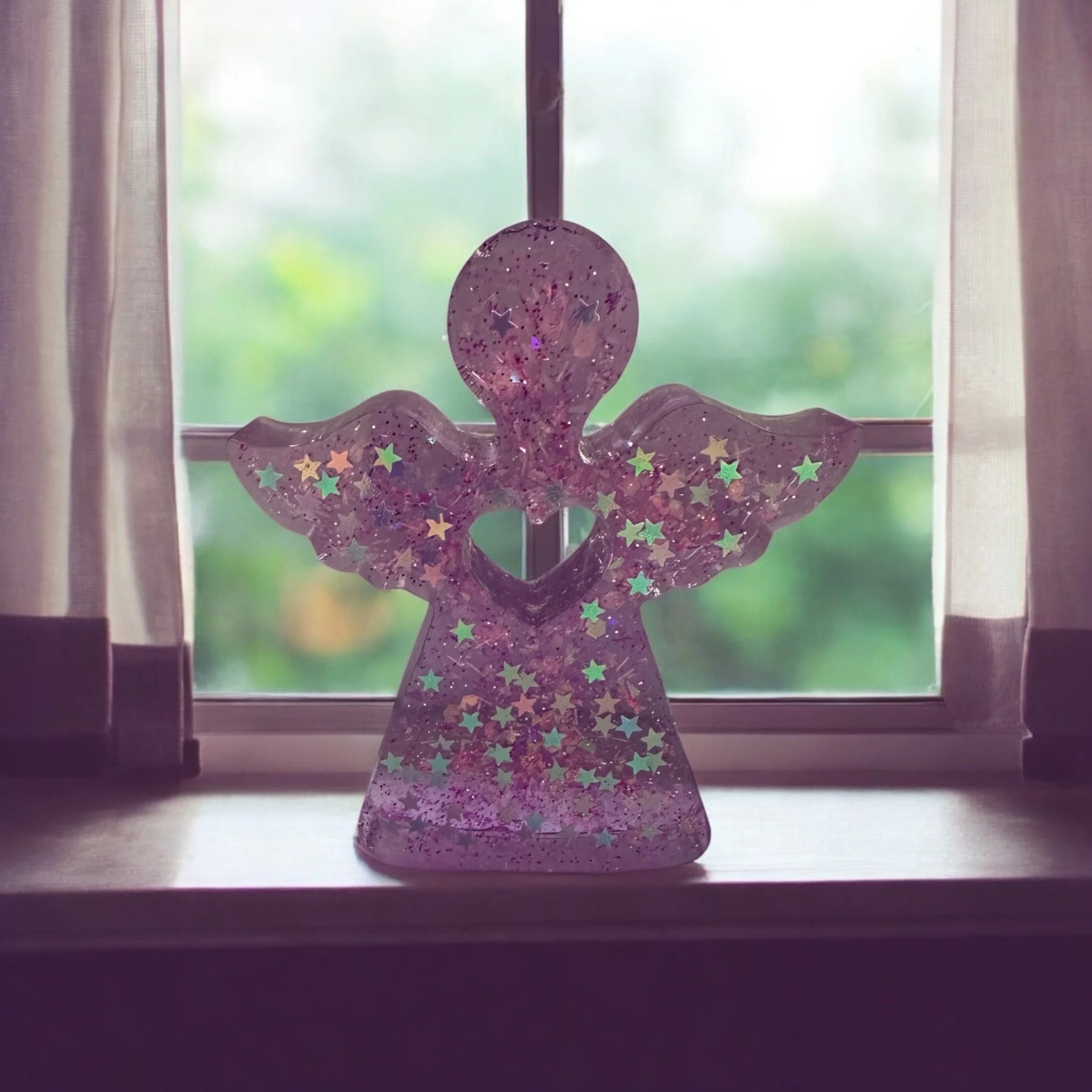 A decorative angel-shaped figure with star-shaped glitter inside, placed on a windowsill with sheer curtains. Outside the window, green foliage is visible.