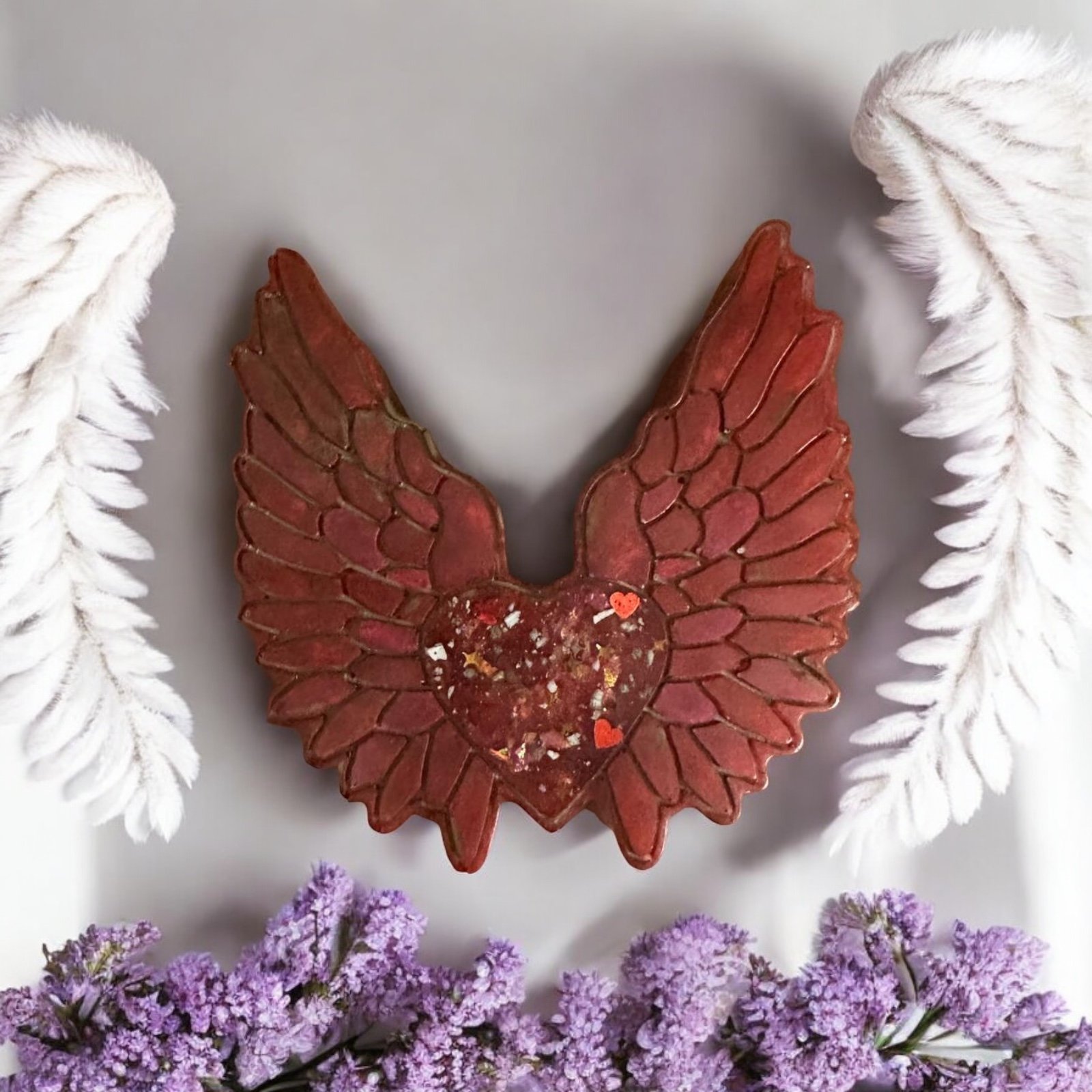 Decorative wall hanging of a heart with wings, painted red with heart-shaped accents, surrounded by white feather-like motifs and purple flowers at the bottom.