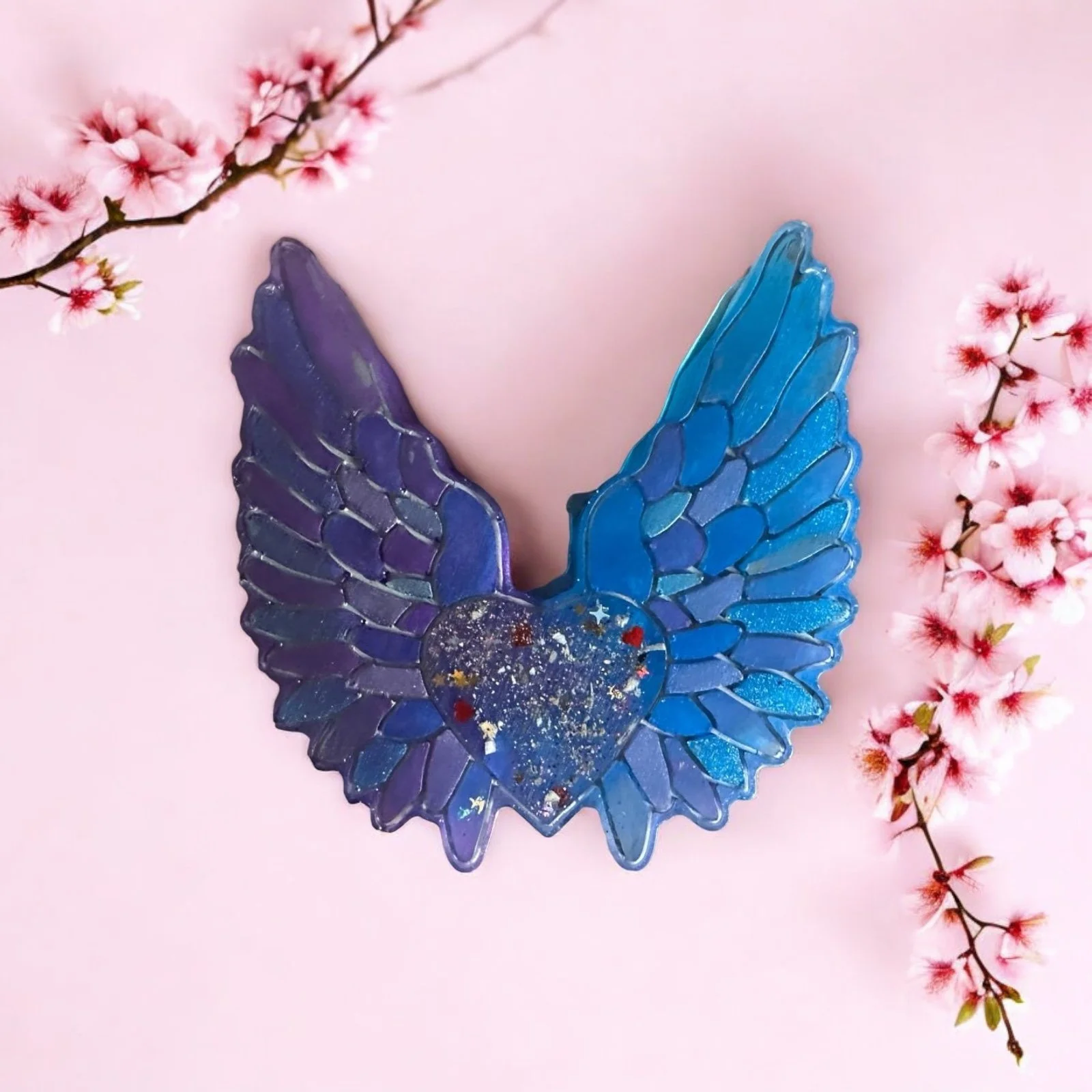 A decorative glass piece shaped like a heart with wings, featuring blue and purple hues and gold accents, placed on a pink background with pink cherry blossom branches on both sides.