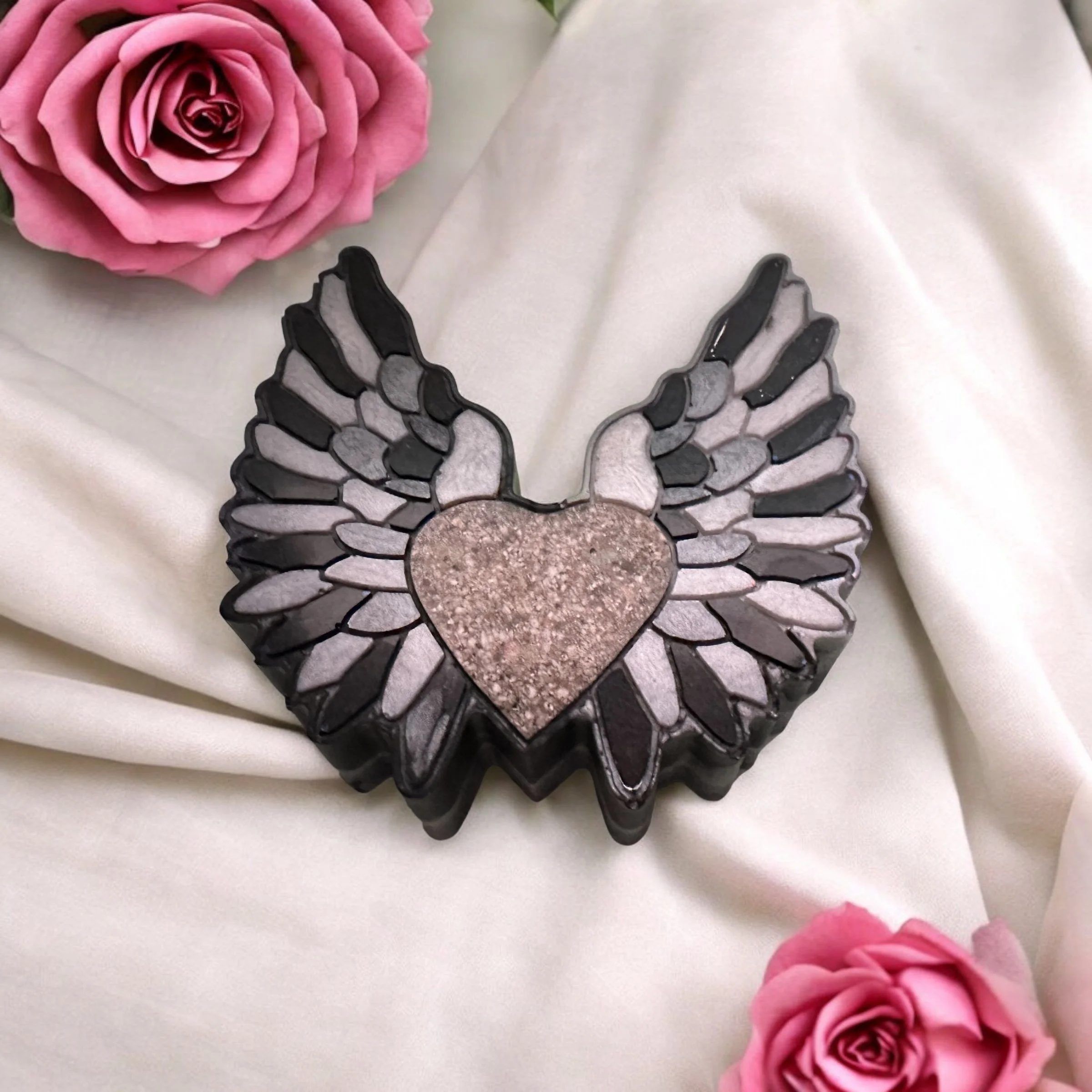 Decorative piece with a heart center and black and silver wings; surrounded by pink roses and white fabric.