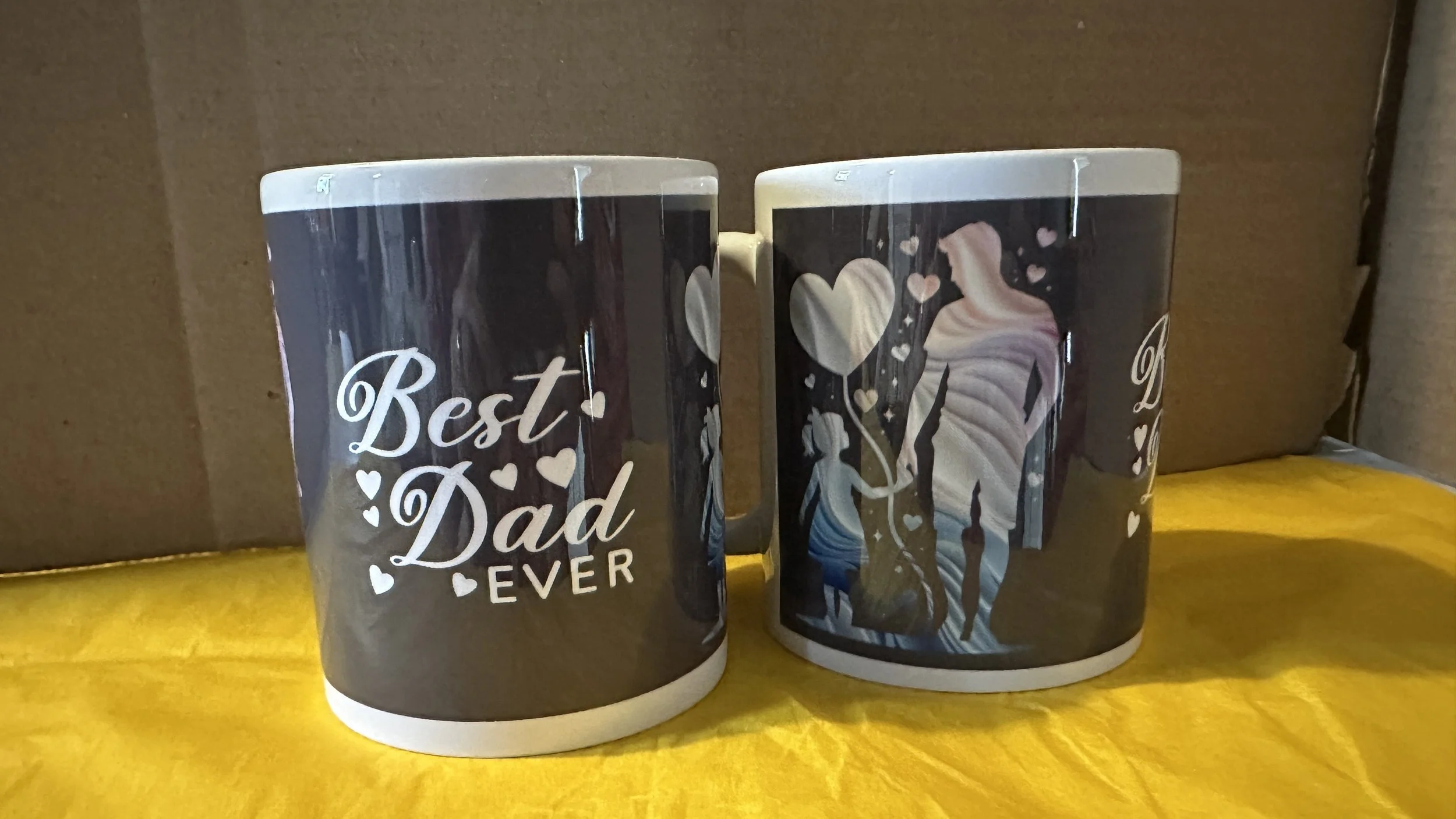 Two coffee mugs with black and white designs; one says 'Best Dad Ever' and the other shows a silhouette of a couple holding balloons, surrounded by hearts.