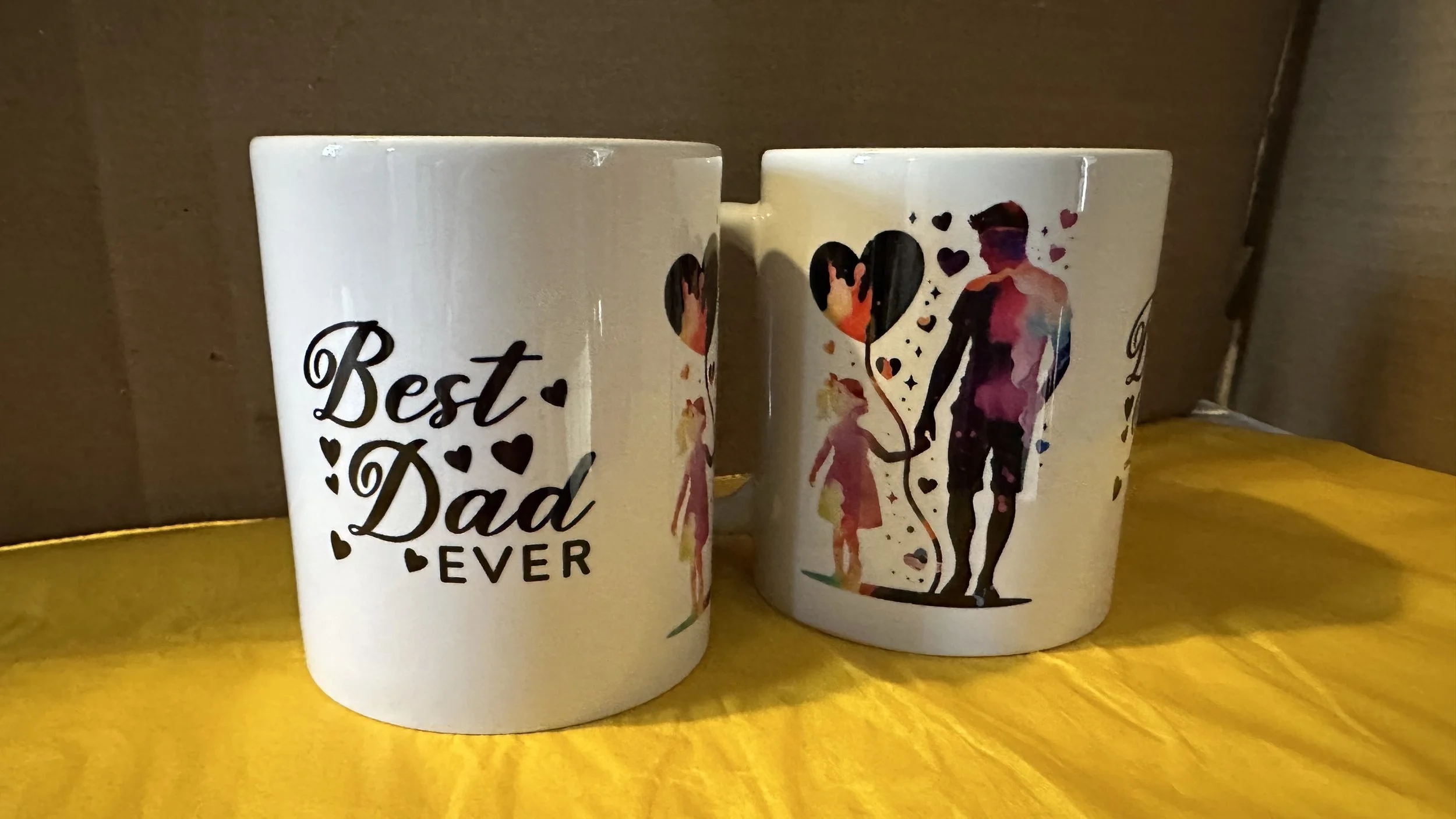 Two white ceramic mugs on a yellow surface. One mug has the text 'Best Dad Ever' with small black hearts, and the other mug shows an artistic watercolor-style silhouette of a man and a young girl holding heart-shaped balloons.