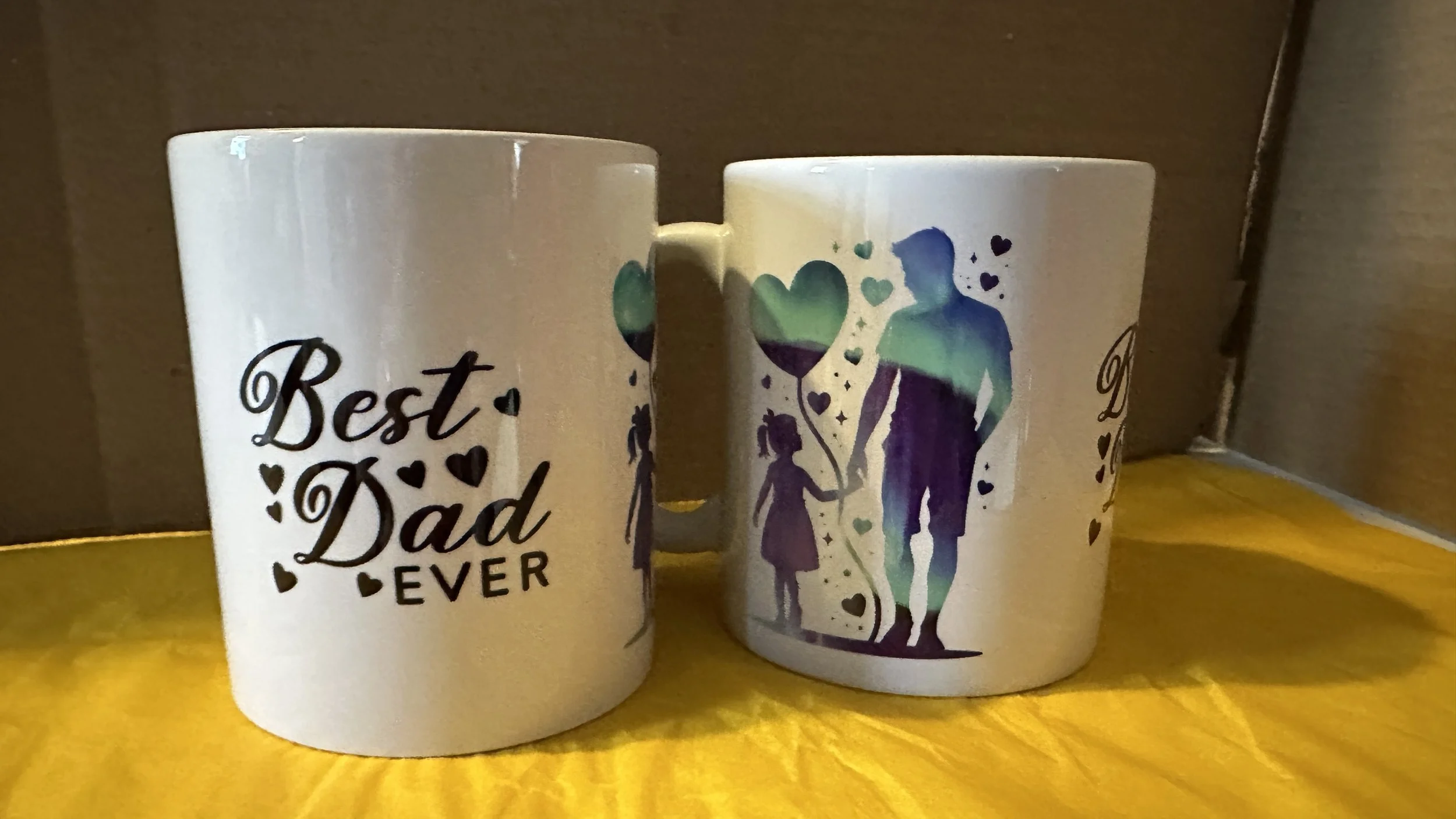 Two white ceramic mugs with black and colorful printed designs. The mug on the left has the text "Best Dad Ever" surrounded by small hearts. The mug on the right features a silhouette of a man and a girl holding hands, with additional hearts and balloon illustrations, against a background of blue and green watercolor effects.