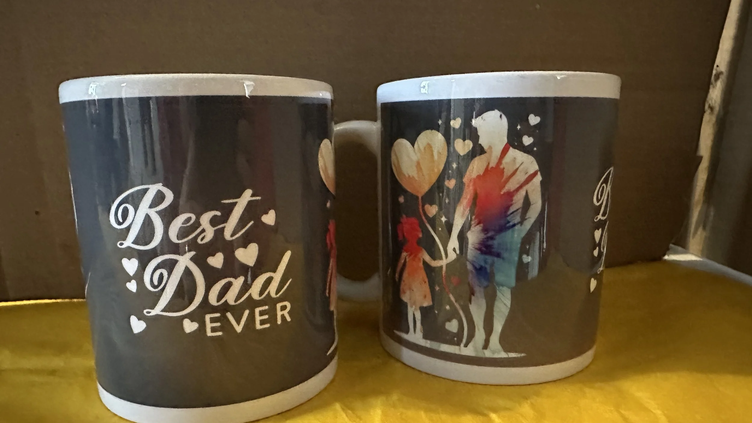 Two mugs with a black background and white text that reads 'Best Dad Ever', with small hearts. The mugs also feature a colorful illustration of a father and child holding balloons, with the father figure shirt painted in a rainbow watercolor style.