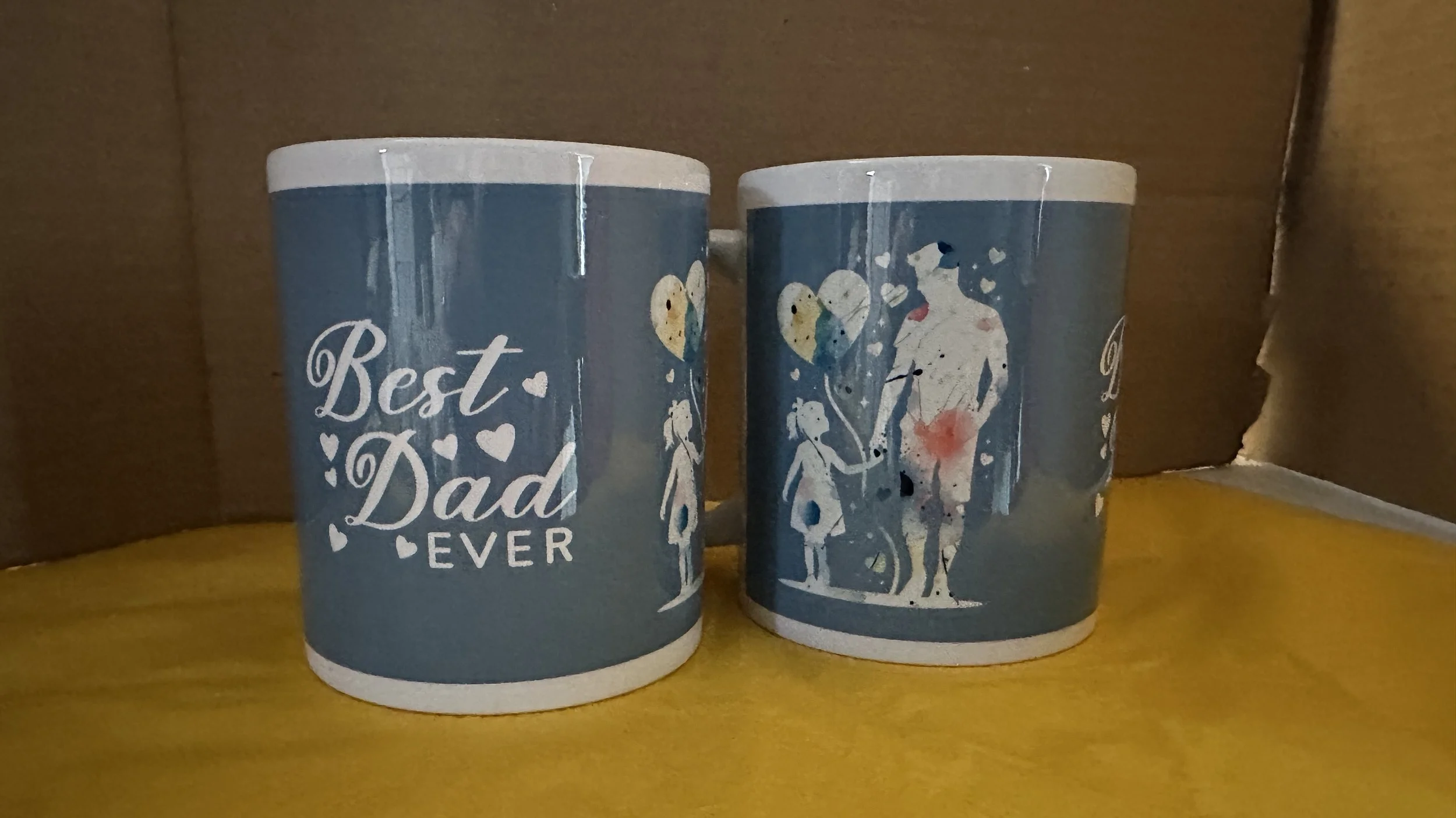 Two blue coffee mugs, one with the text 'Best Dad Ever' and heart designs, the other with a graphic of a man holding hands with two children, against a brown background.
