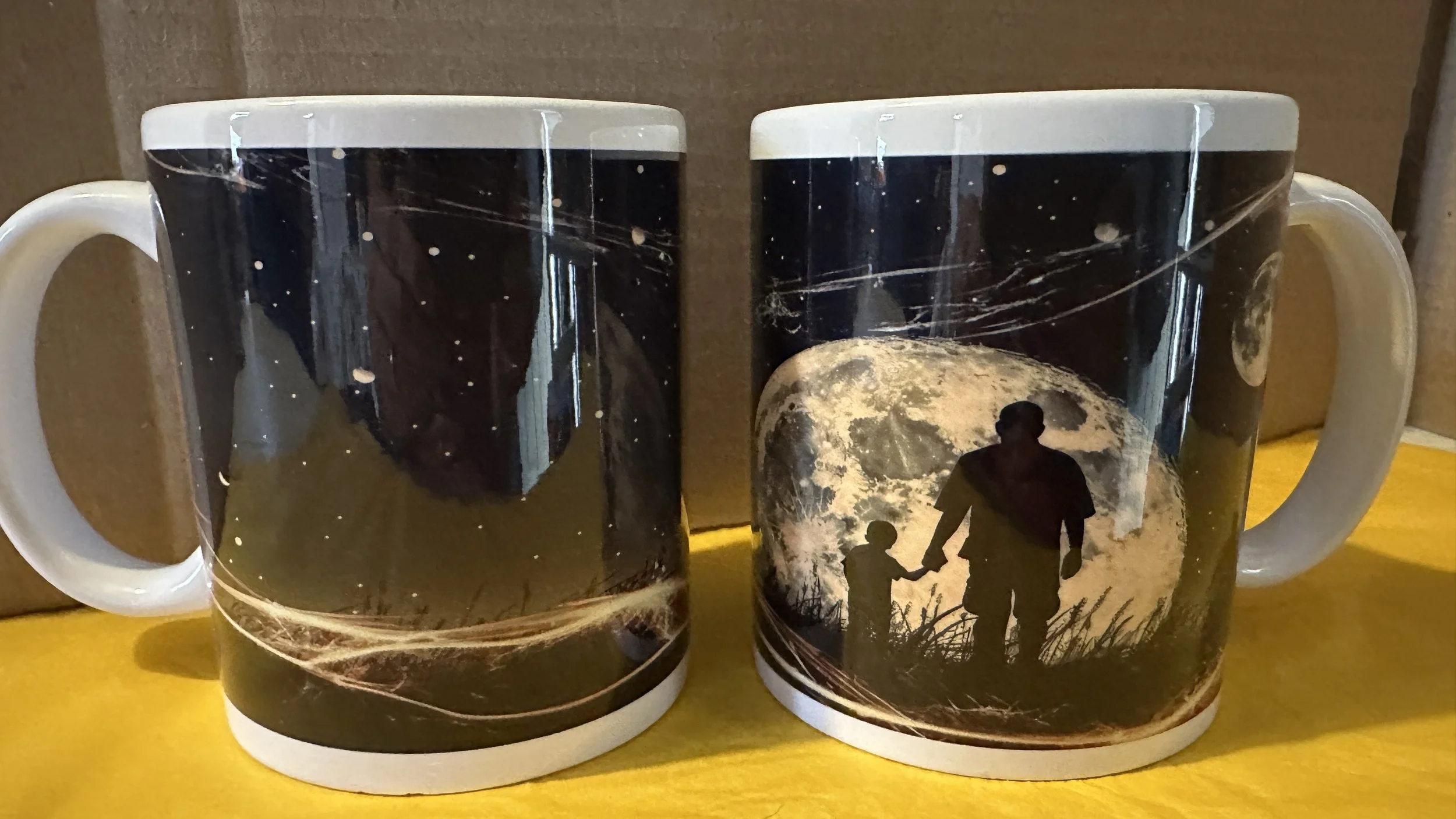Two coffee mugs with a silhouette of an adult and child holding hands against a large full moon, with space and star patterns in the background.