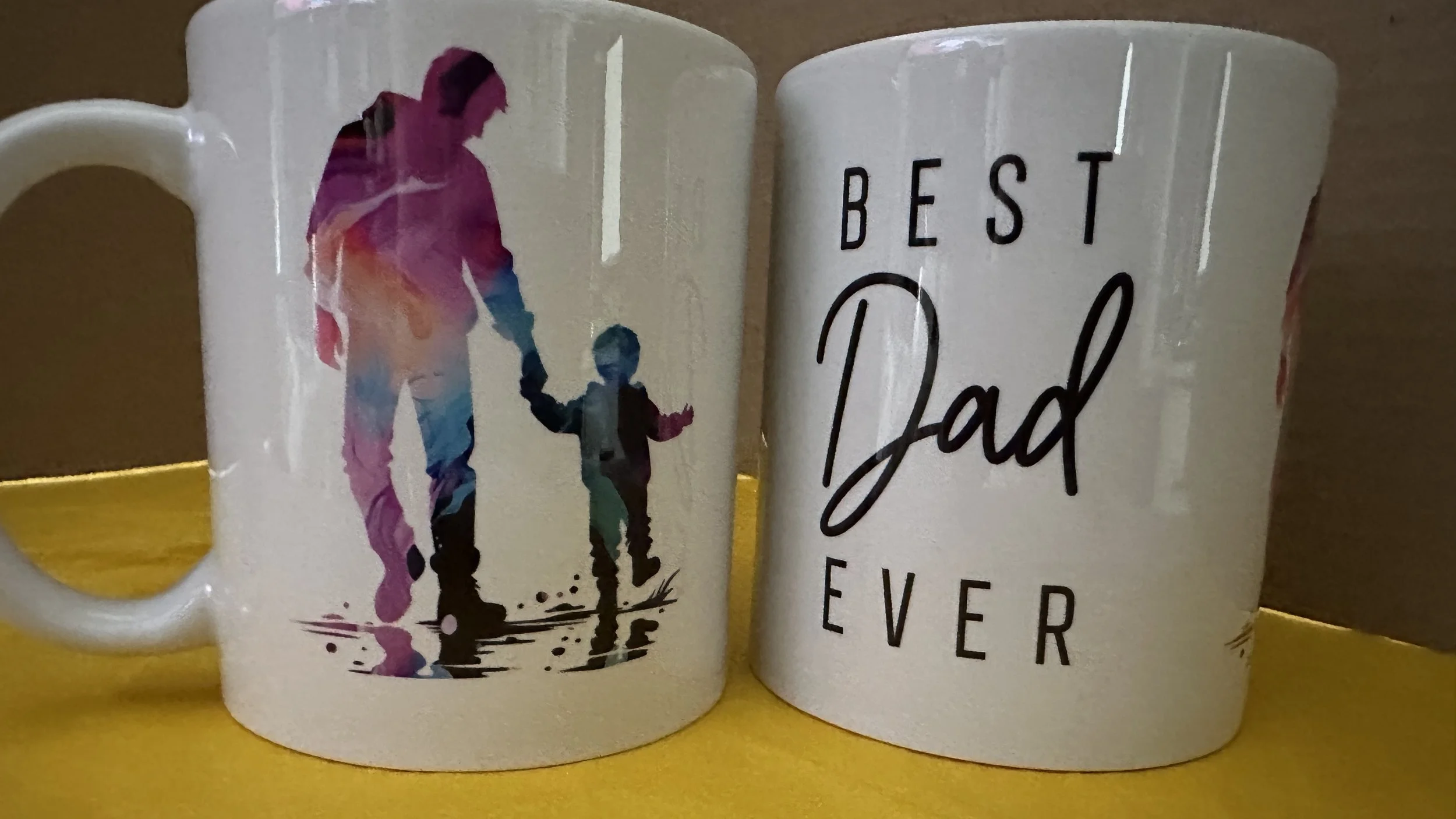 Two white ceramic mugs; one features an artistic colorful silhouette of an adult and child holding hands, the other has the words "Best Dad Ever" in black lettering.