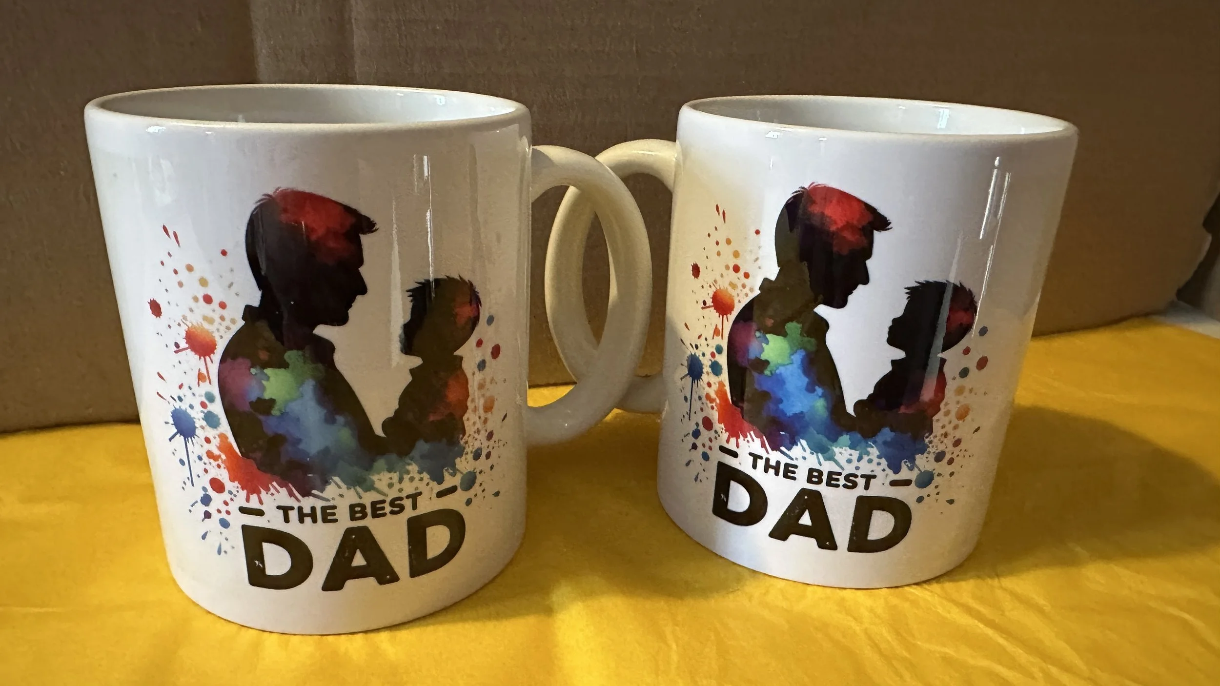 Two white coffee mugs with silhouettes of a father and child facing each other, surrounded by colorful paint splashes, and the text "The Best Dad" printed at the bottom.