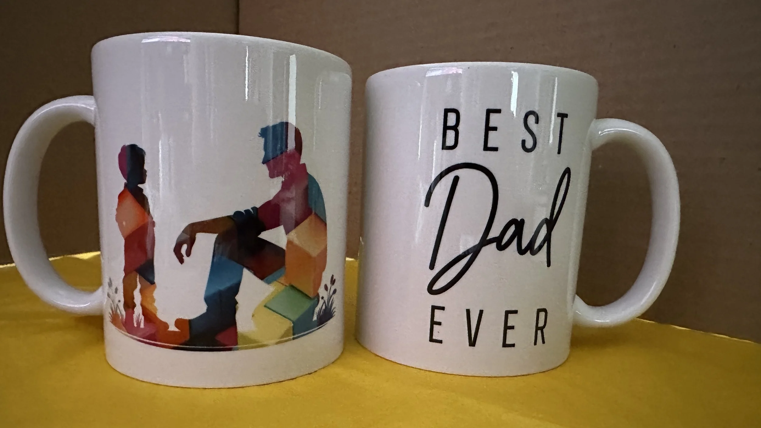 Two white ceramic coffee mugs on a yellow surface against a wooden background. One mug features colorful silhouettes of a father and child with books, and the other mug has the text "Best Dad Ever" in black and cursive.