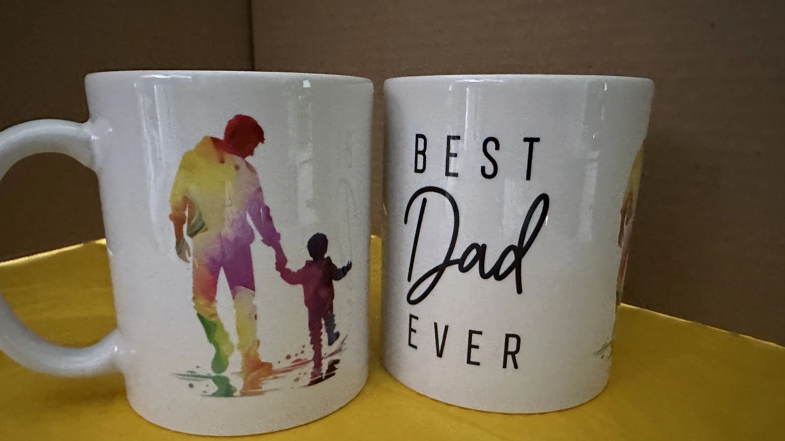 A set of two white coffee mugs, one featuring a colorful watercolor silhouette of a father walking with a child, and the other with the text "Best Dad Ever" in black lettering.