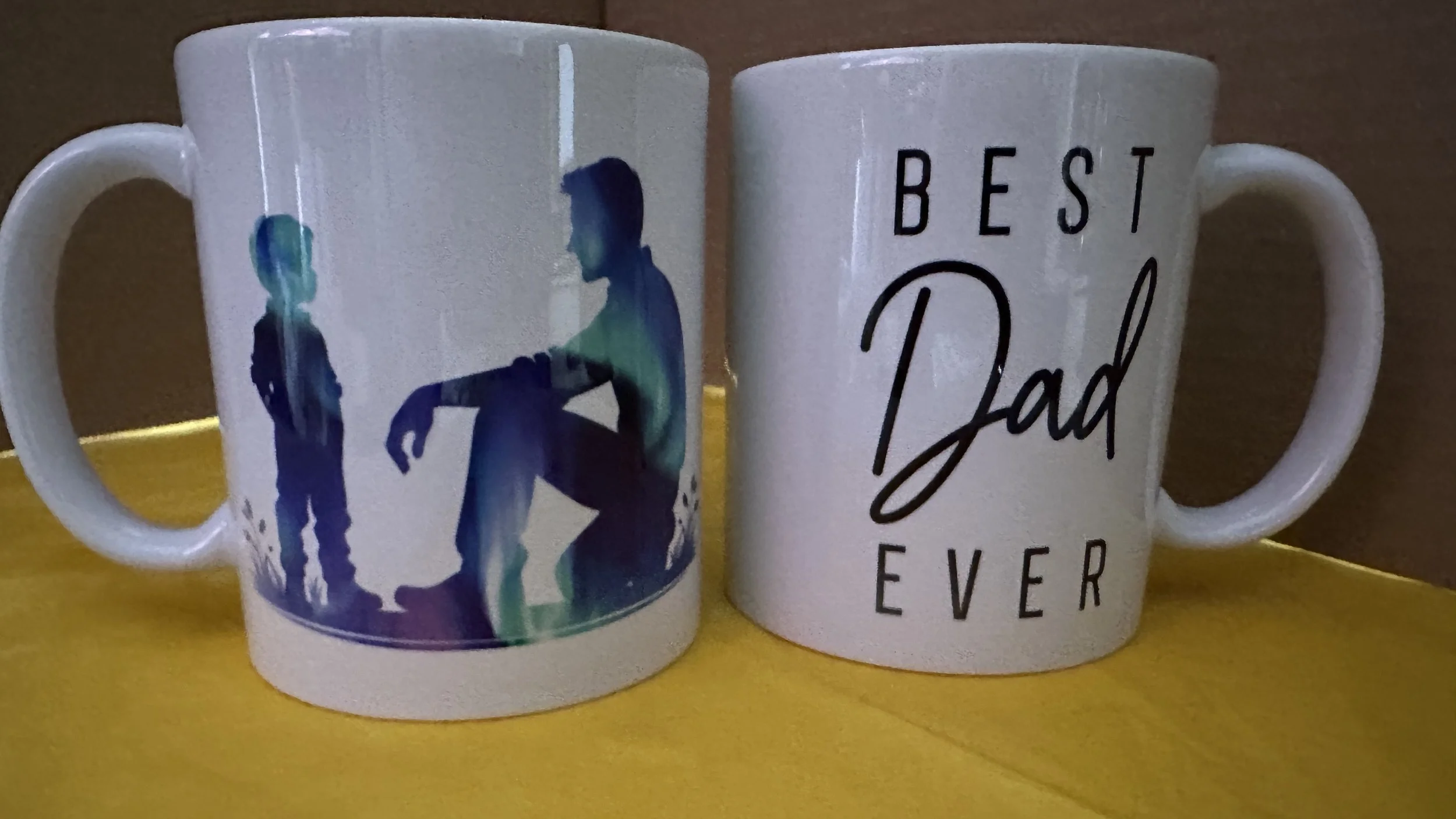 Two white coffee mugs with silhouette graphic and text. The left mug features a silhouette of a child and an adult with the adult kneeling and extending an arm to the child. The right mug has the phrase "Best Dad Ever" written in black script and capital letters.
