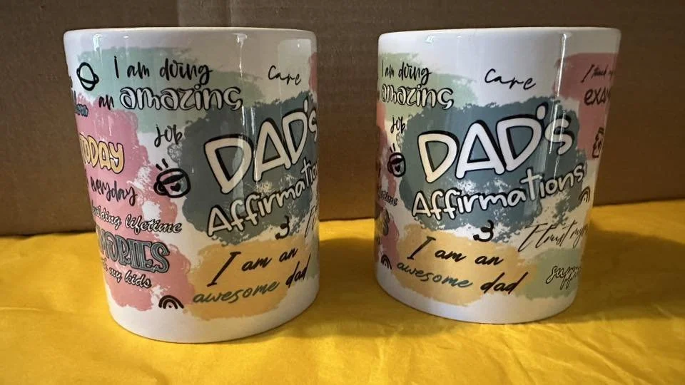 Two white ceramic mugs with colorful text and designs celebrating fathers, including phrases like "DAD's Affirmations," "I am doing an amazing job," and "I am an awesome dad," placed on a yellow surface with a cardboard box background.
