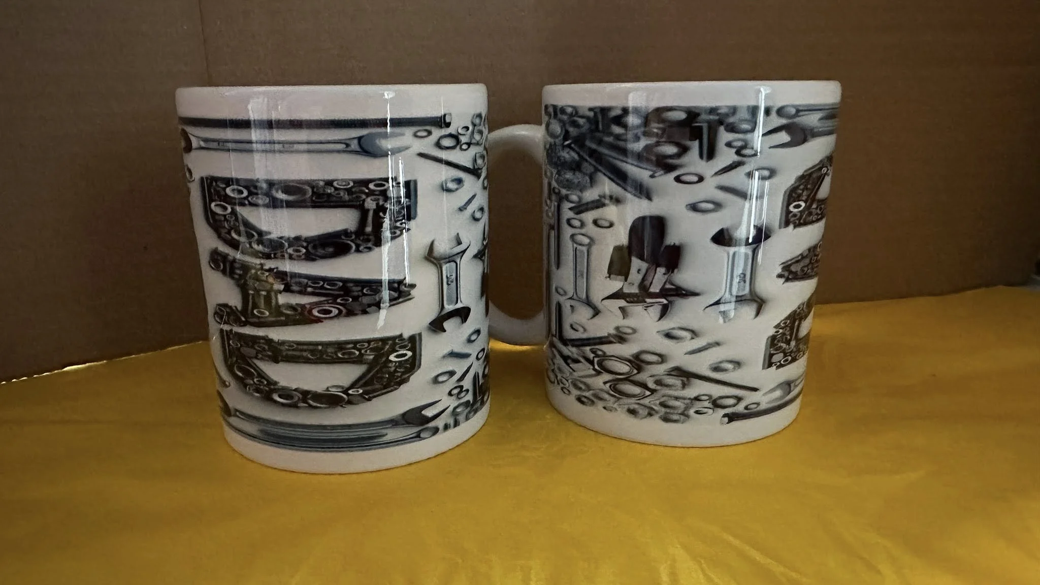Two white ceramic mugs with black and gray mechanical and tool illustrations, placed on a yellow surface against a brown background.