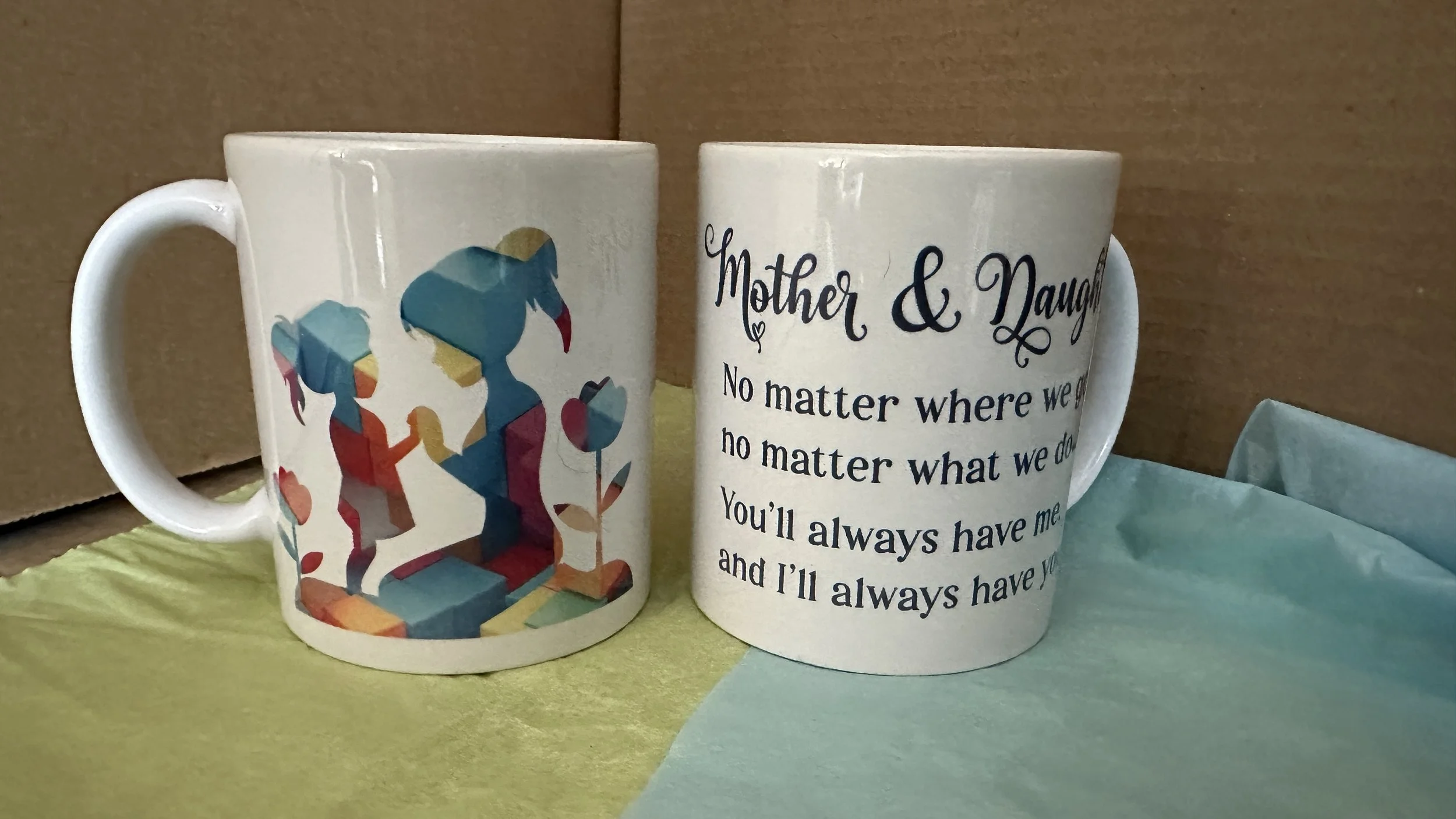 Two white ceramic mugs with colorful and printed designs, placed on a yellow and green surface against a brown cardboard background. One mug features a pixelated image of a mother bird and her chick with a red beak, and the other mug has the text: 'Mother & Daughter. No matter where we go or what we do, you'll always have me and I'll always have you.'
