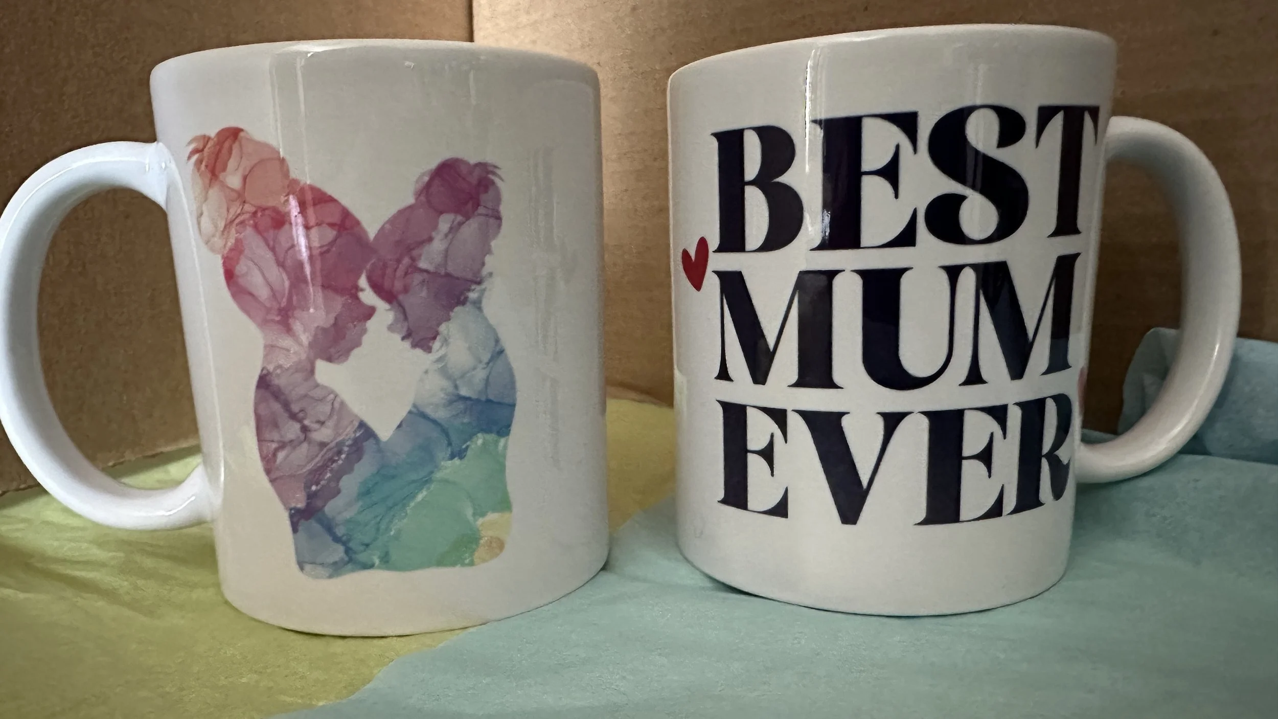 Two ceramic mugs, one with an abstract watercolor image of a woman and the other with the text 'Best Mum Ever' and a small red heart.