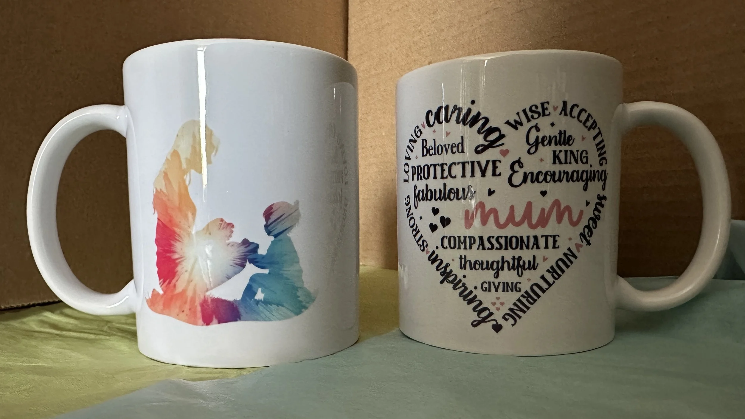 Two white mugs on a table, one featuring an illustration of a child and a woman holding hands with colorful watercolor effects, and the other with a heart-shaped design filled with words describing a mother, such as 'loving,' 'protective,' 'fabulous,' and 'mum' in various fonts and colors.