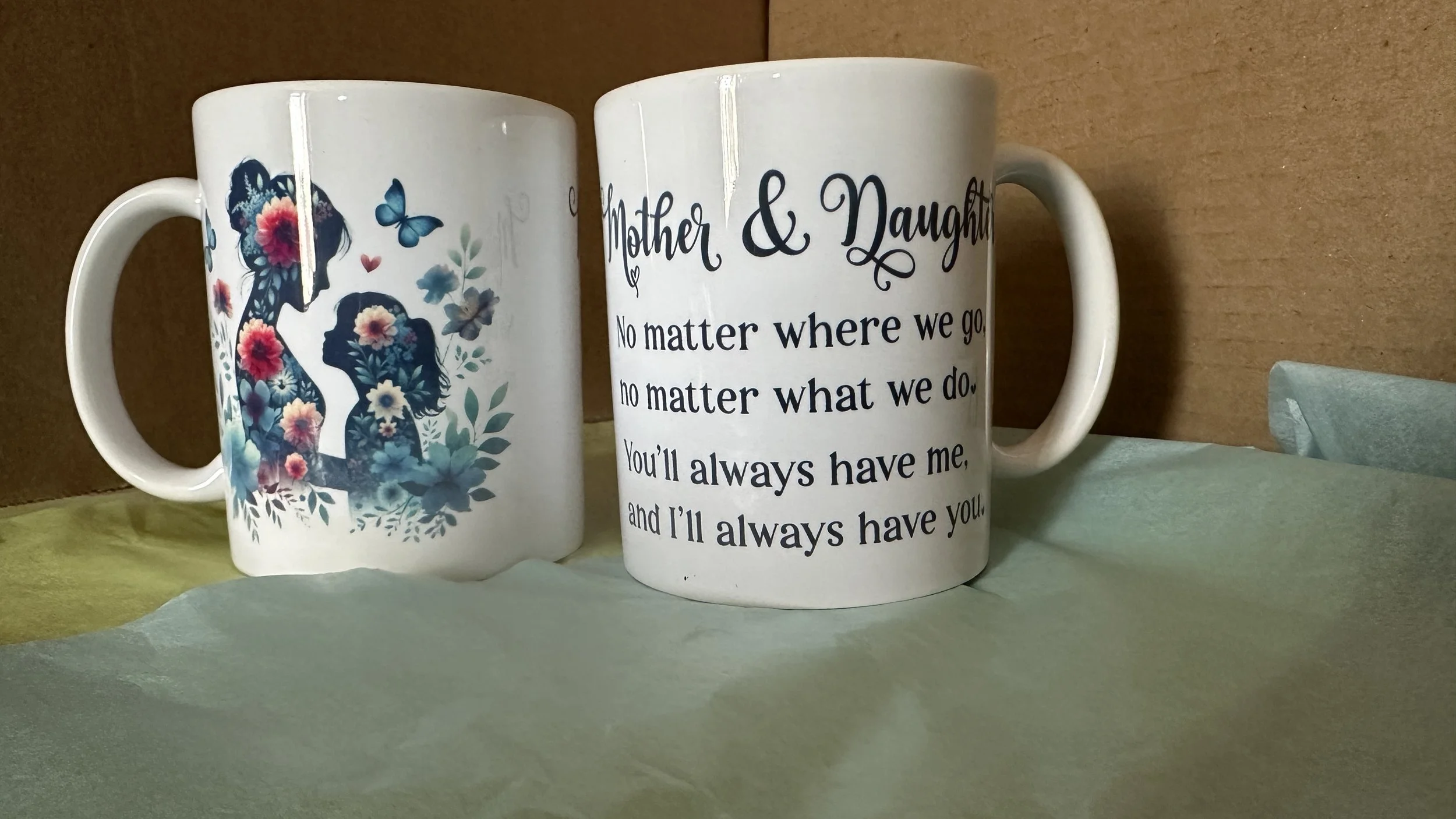 Two white ceramic mugs, one with a floral silhouette of a woman and girl with flowers and butterflies, and the other with a quote about mothers and daughters.