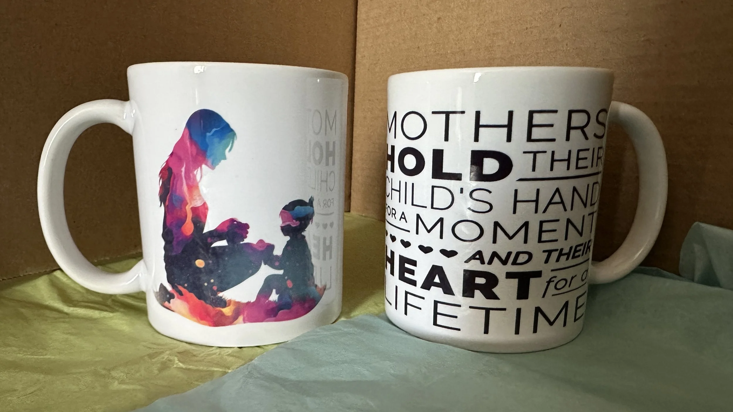 Two white ceramic mugs with different designs, placed side by side. The mug on the left features a colorful, watercolor-style silhouette of a woman and a child sitting and holding hands. The mug on the right has a black and white typographic design with words about mothers holding children's hands for a lifetime.