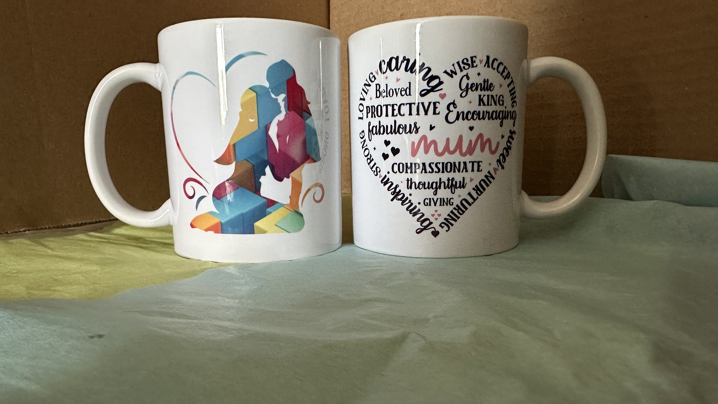 Two white ceramic mugs, one featuring a colorful abstract illustration of a mother and child, and the other with a heart-shaped arrangement of words describing a mother, including loving, caring, protective, and compassionate, with the word 'mum' in the center.