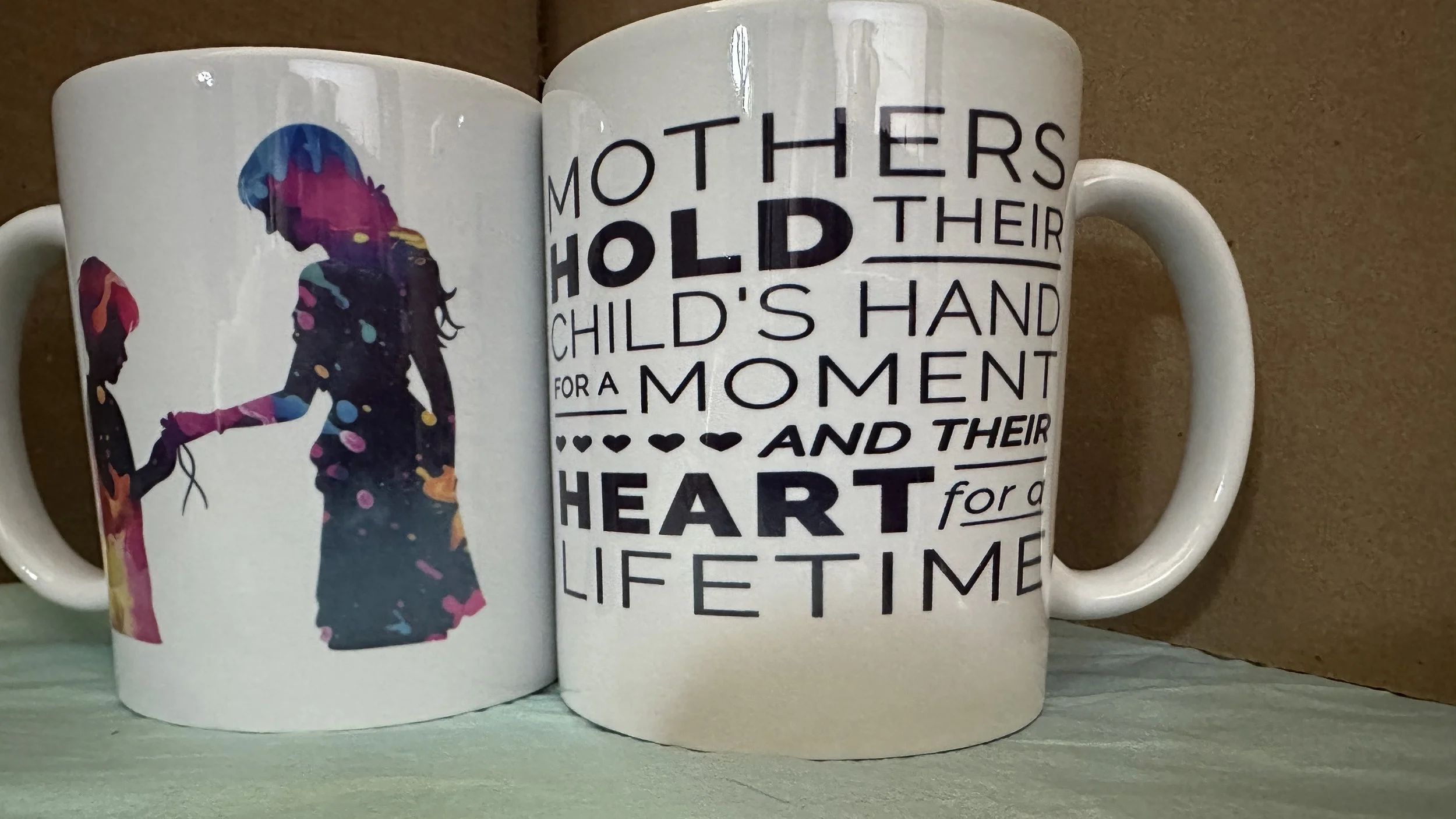 Two ceramic mugs, one featuring silhouettes of a child and an adult holding hands, the other with a quote about mothers and children in black text.
