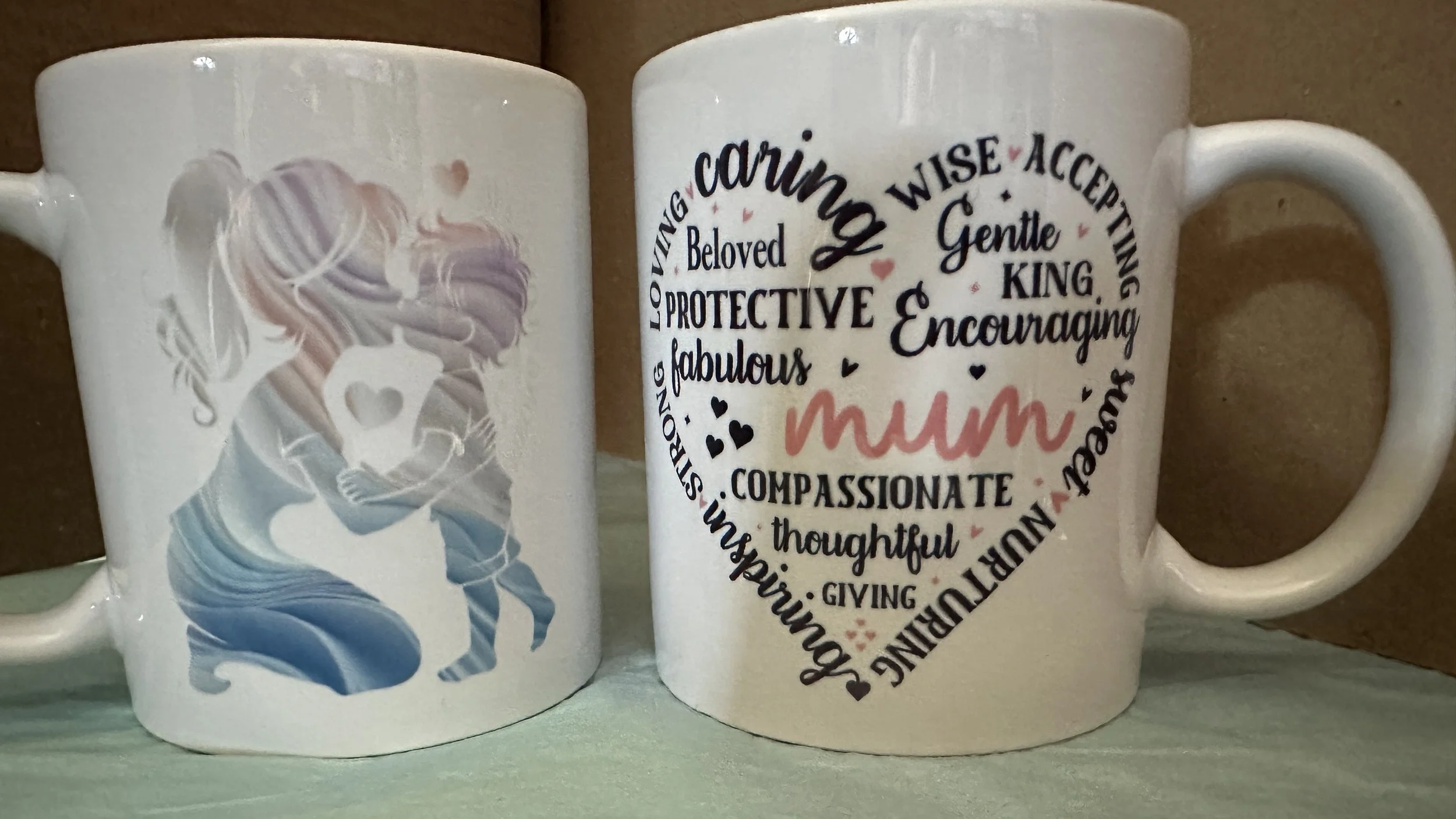 Two white ceramic mugs on a surface. The mug on the left has a design of a girl with long hair holding a heart-shaped lollipop. The mug on the right has a word cloud in the shape of a heart with words like caring, mom, nurturing, wise, gentle, king, encouraging, compassionate, thoughtful, loving, protective, fabulous, and accepting.