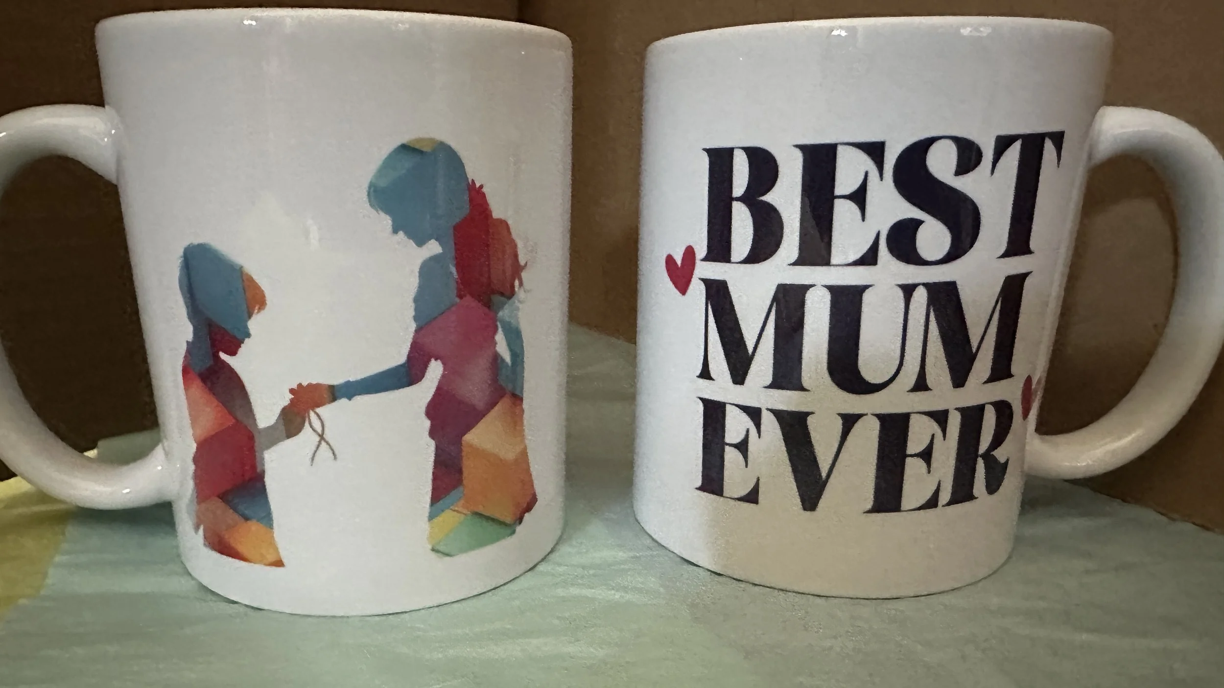 Two white coffee mugs, one with a colorful illustration of a woman handing a gift to a girl, the other with black text that says "BEST MUM EVER" and small red hearts.