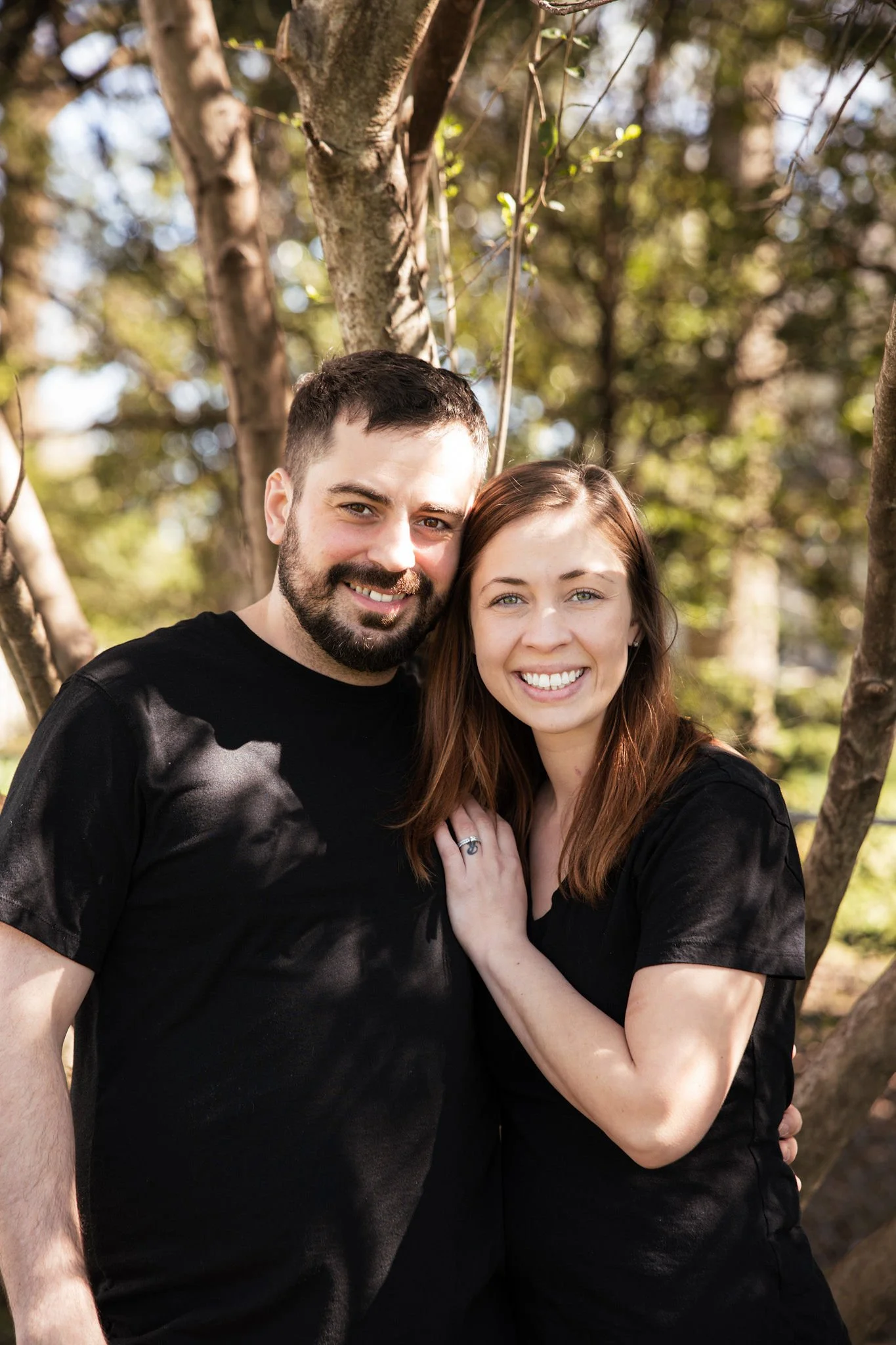 A smiling couple standing close together outdoors by a tree, with the man on the left and the woman on the right, both wearing black shirts.