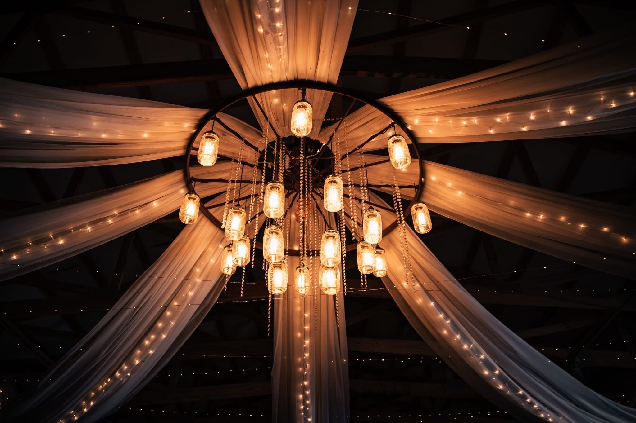 A chandelier with multiple warm-colored lights hangs from a ceiling decorated with draped fabric and string lights, creating a warm, elegant ambiance.