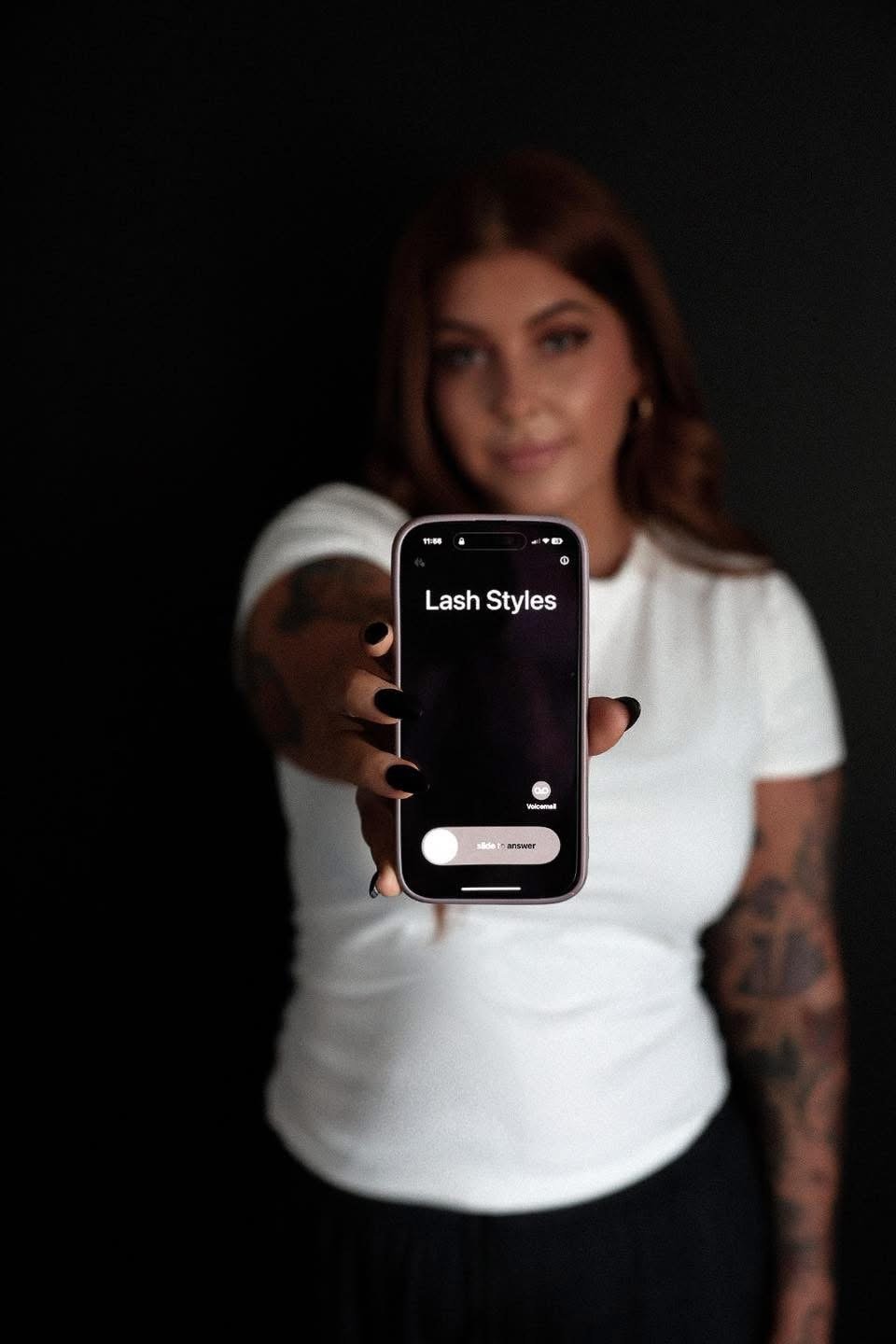 A woman with brown hair, tattoos on her arm, wearing a white t-shirt, holds a smartphone displaying a screen titled 'Lash Styles'. She has black nail polish and is posing against a dark background.